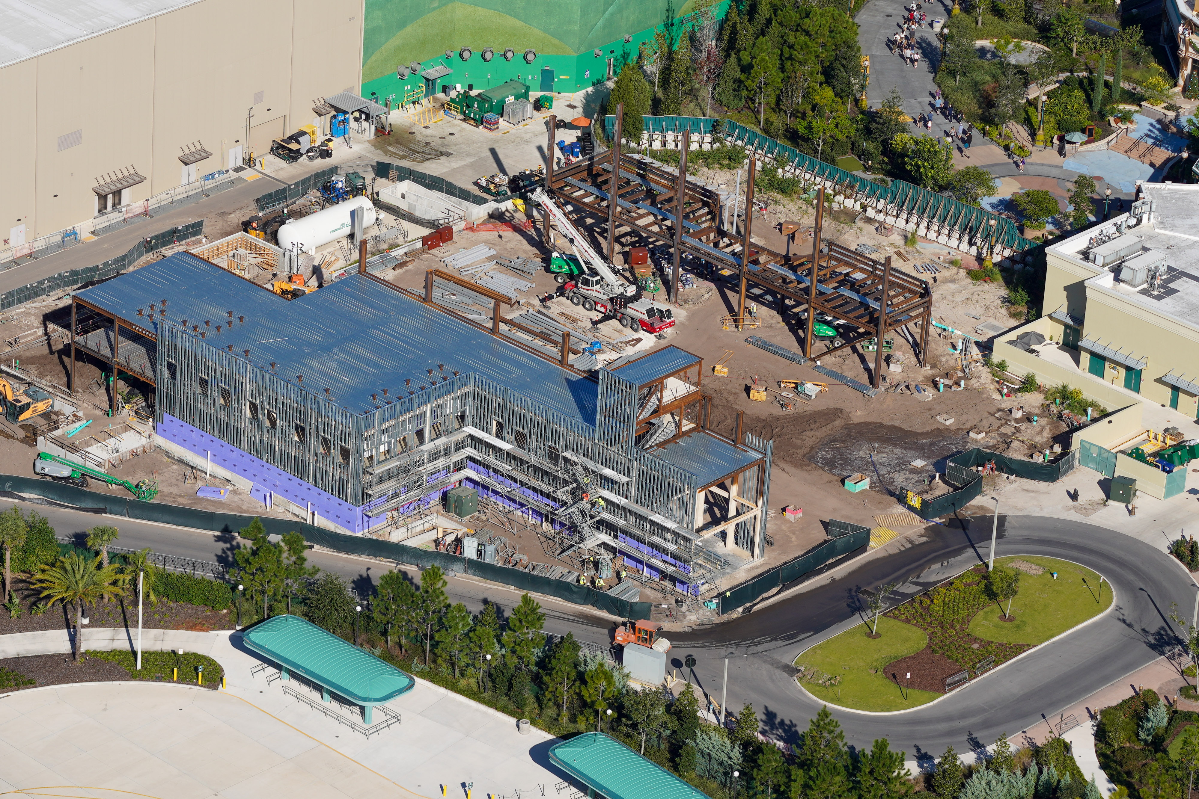 Aerial view of a two-story building under construction with metal roof decking, steel stud walls and purple sheathing along its base, beside a bare steel frame, a crane, and a curving backstage road