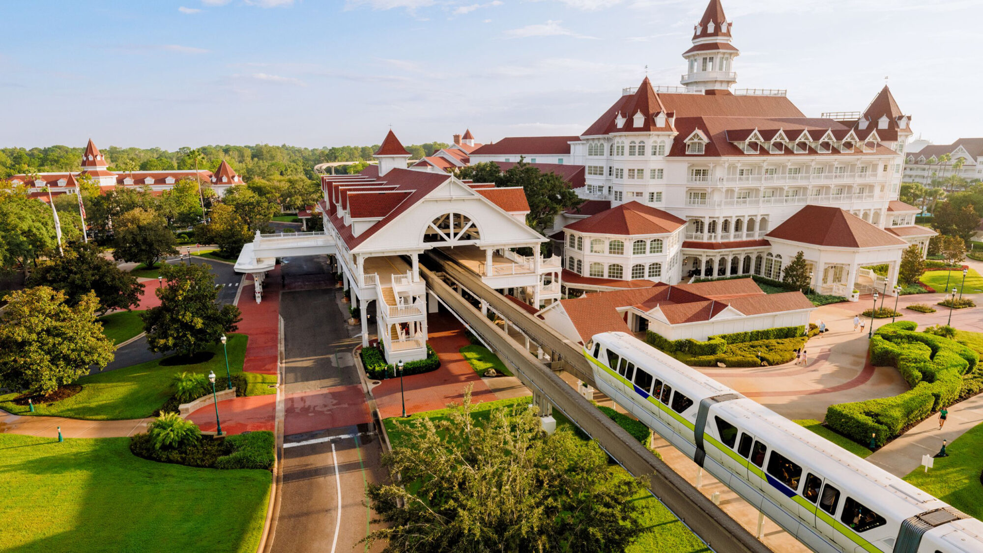 Aerial view of Disney's Grand Floridian Resort and Spa, white Victorian buildings with red roofs, with a monorail train with a lime green stripe passing on the beam in the foreground