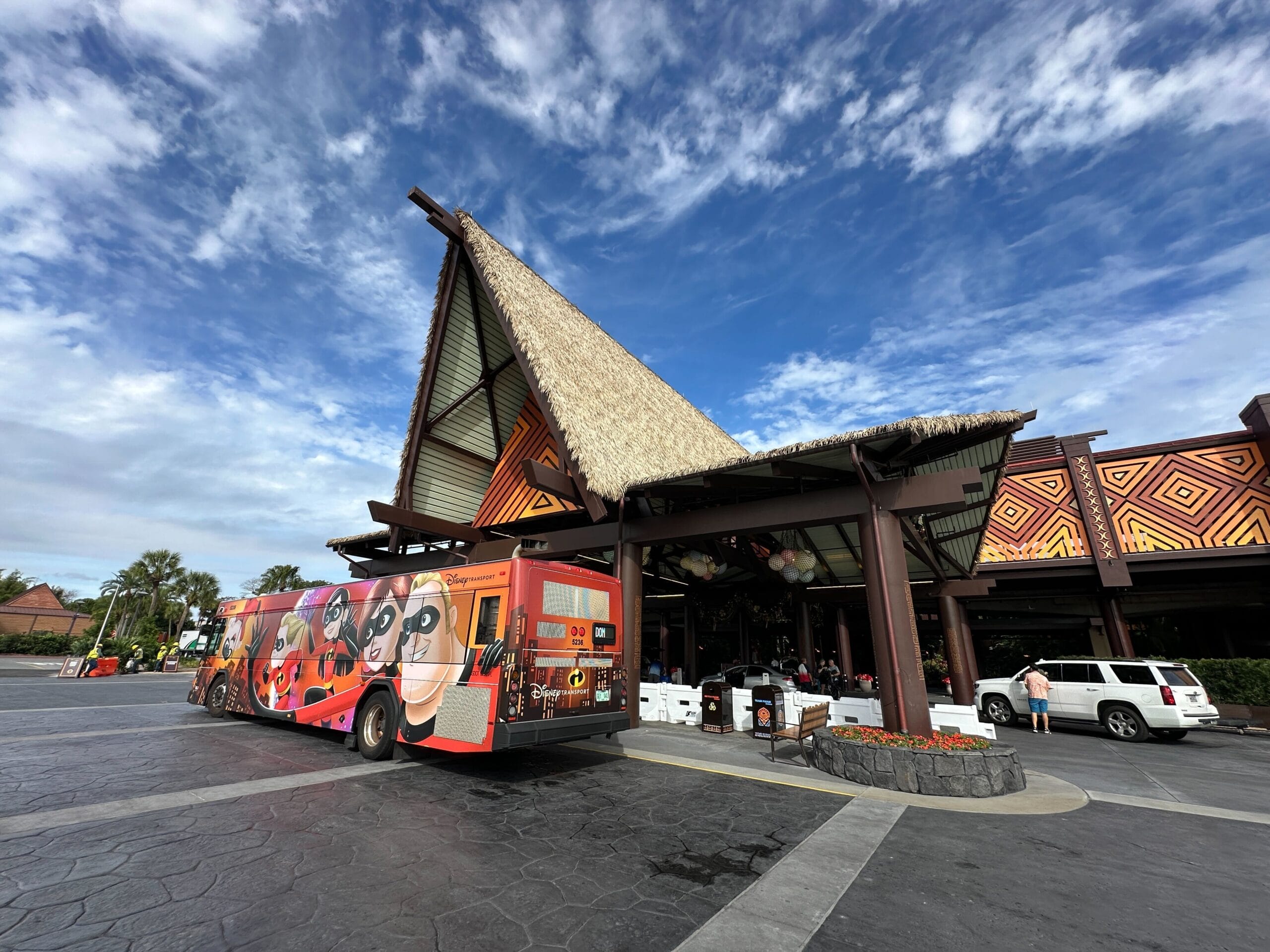 The thatched, steeply peaked porte-cochere entrance of Disney's Polynesian Village Resort under a blue sky, with an orange Incredibles-wrapped Disney Transport bus parked in front