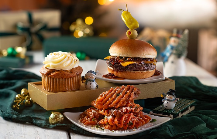 A cheeseburger topped with onion jam and a skewered pepper, a frosted cupcake on a gold tray, and a plate of seasoned waffle fries with grated cheese, with snowman figurines on a green cloth