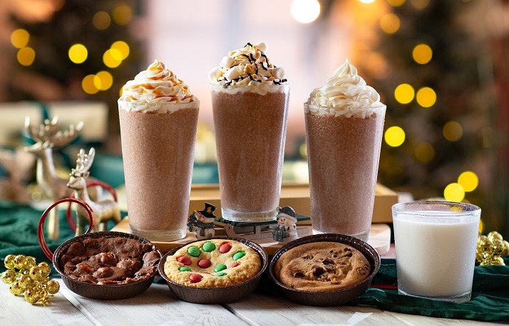 Three tall frozen drinks topped with whipped cream, two with caramel or chocolate drizzle, behind three deep dish cookies in small tins and a glass of milk, with reindeer figurines at the left and Christmas lights behind