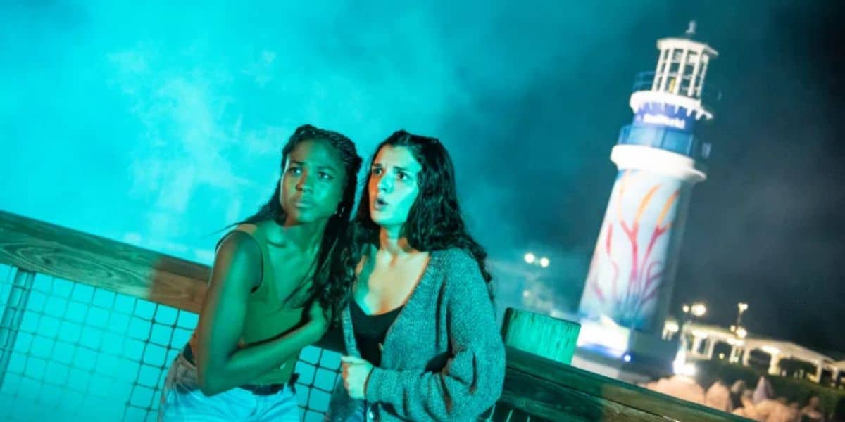 Two guests stand at a wooden railing at night during Howl-O-Scream, lit in teal light with fog billowing behind them, both looking off camera and the woman on the right wide-eyed and open-mouthed. An illuminated lighthouse rises in the background against a dark sky.