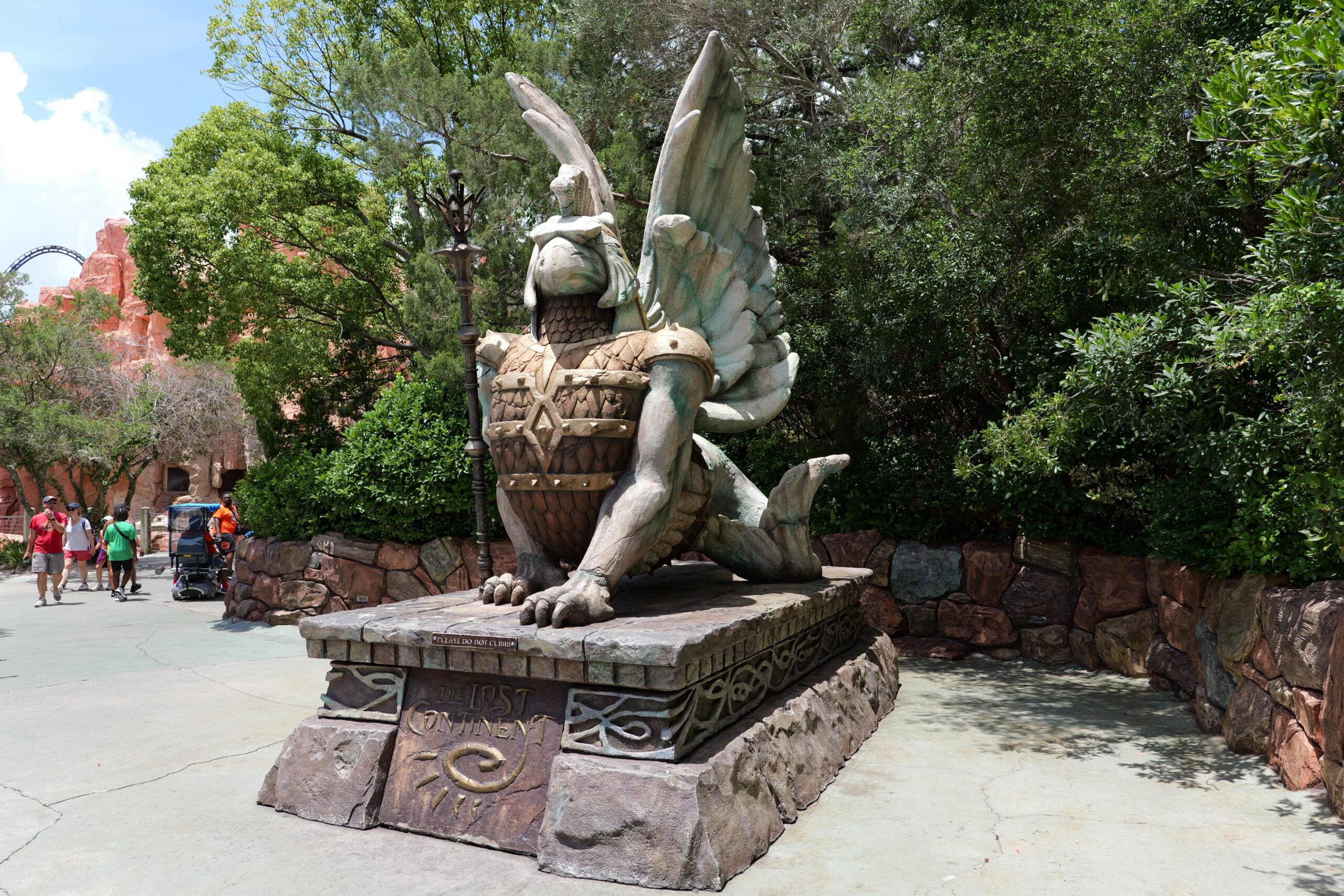 A weathered green and stone griffin statue perched on a carved plinth reading The Lost Continent, at the entrance to the land at Universal Islands of Adventure, with red rockwork and a roller coaster track visible in the background.