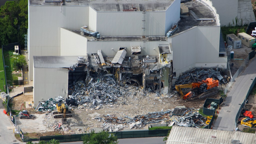 Aerial photo of the Poseidon's Fury show building at Universal Islands of Adventure partially demolished, with an orange excavator working in the wreckage, a second excavator and a compact track loader nearby, and a large pile of twisted metal debris spilling out of the opened side.