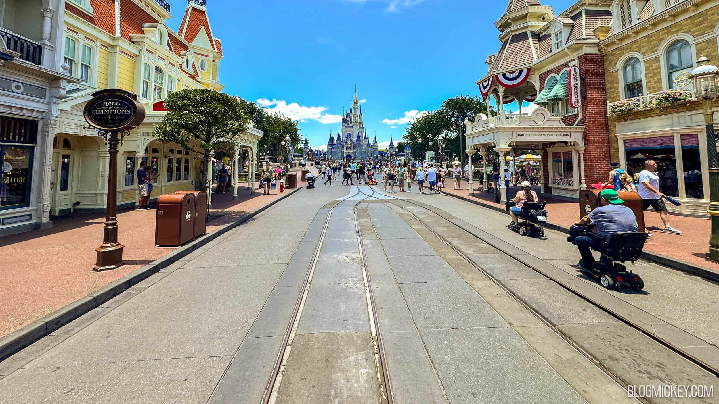 Main Street, U.S.A. at Magic Kingdom on a sunny day, with the trolley tracks running down a roadway clear of guests in the foreground toward Cinderella Castle, while a steady stream of guests fills the sidewalks and the top of the street near the hub.