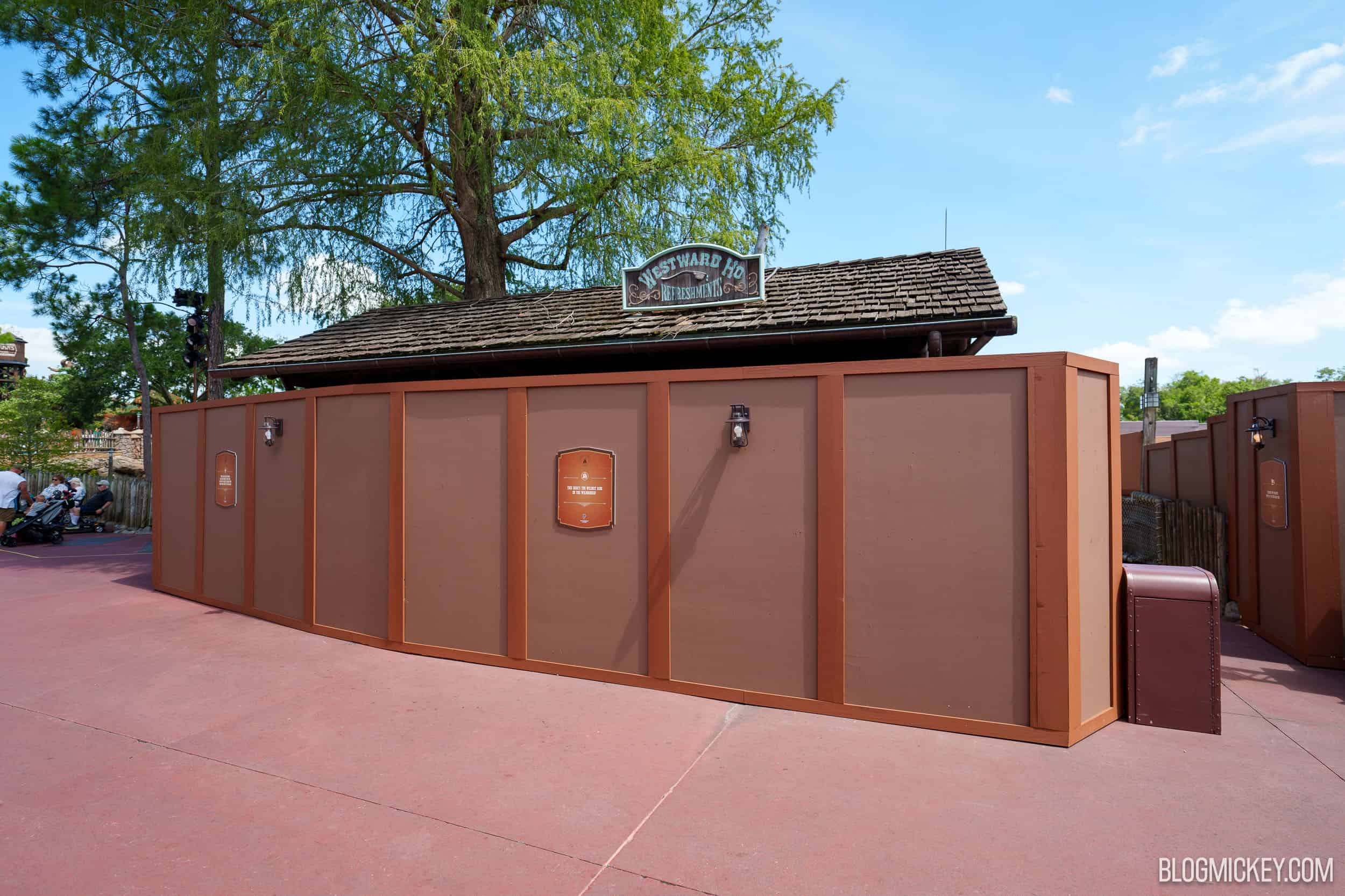 Brown themed construction walls wrapping the Westward Ho Refreshments kiosk in Frontierland at Magic Kingdom, with lantern light fixtures and small themed signs mounted on the walls and the kiosk's wooden shingled roof and Westward Ho Refreshments sign still visible above them, under a blue sky.