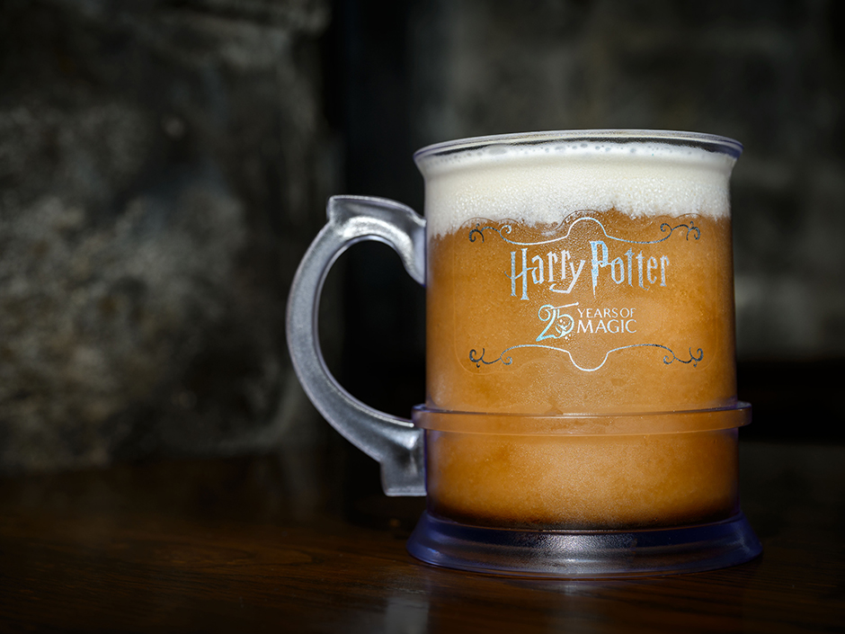 A frosted Butterbeer mug with a thick foam head, printed with the Harry Potter logo above a 25 Years of Magic wordmark framed by scroll flourishes, sitting on a dark wooden table against a stone wall