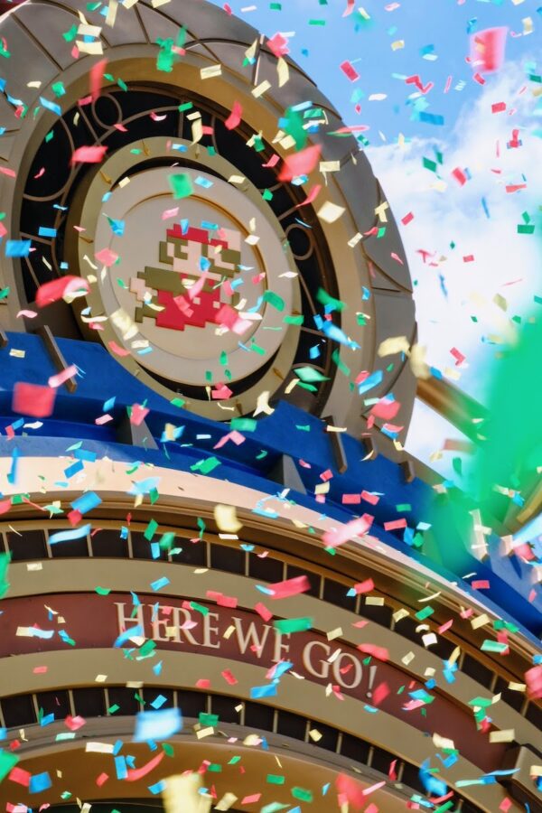 Multicolored confetti filling the air in front of the golden circular Super Nintendo World entrance emblem with a pixel-art Mario figure at its center, above an arched wall reading HERE WE GO!, against a blue sky.