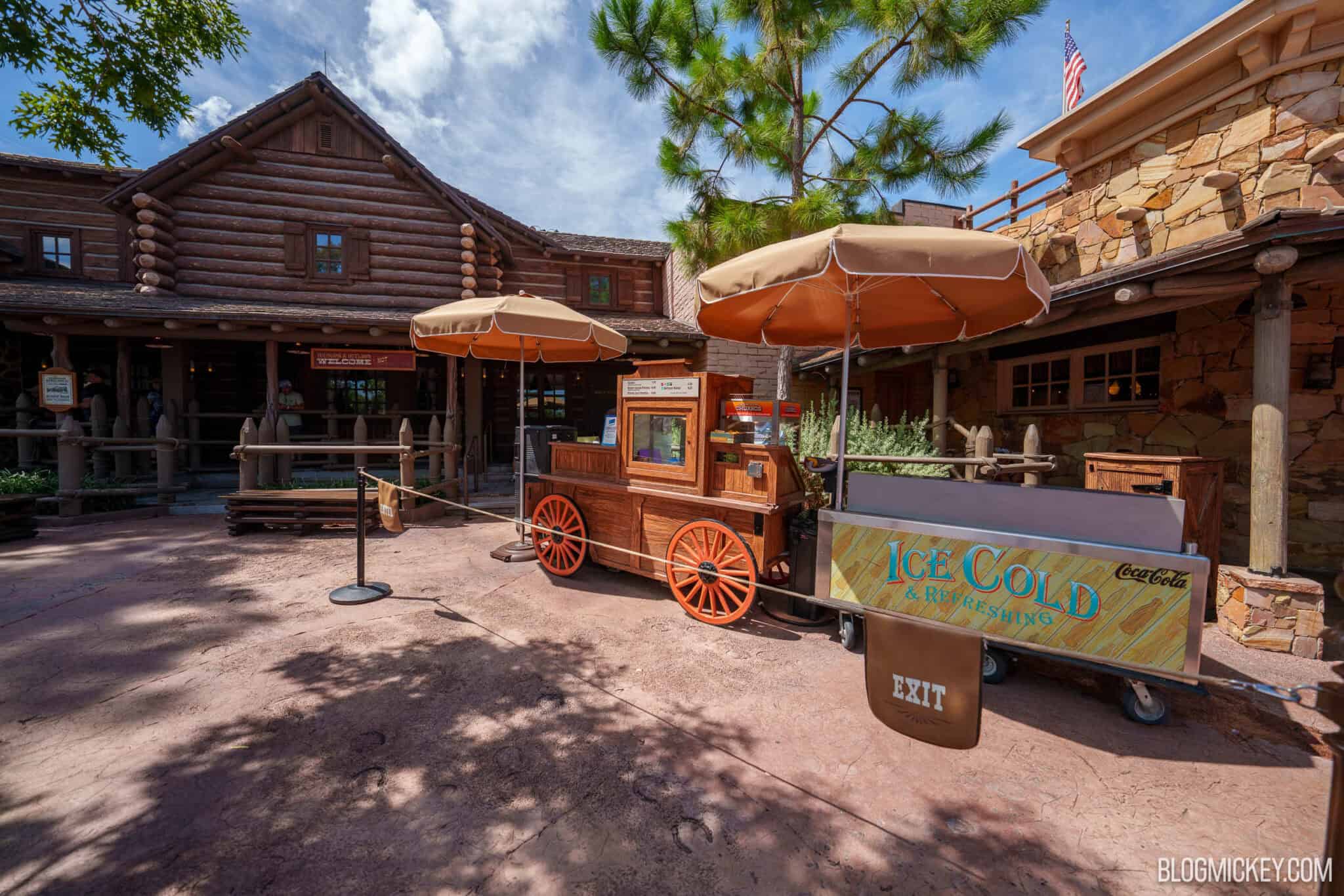 A wooden churro cart with bright orange spoked wheels standing under two umbrellas outside a log building in Frontierland, with a stainless steel Coca-Cola beverage cart parked alongside it, its yellow plank-painted panel lettered Ice Cold and Refreshing.