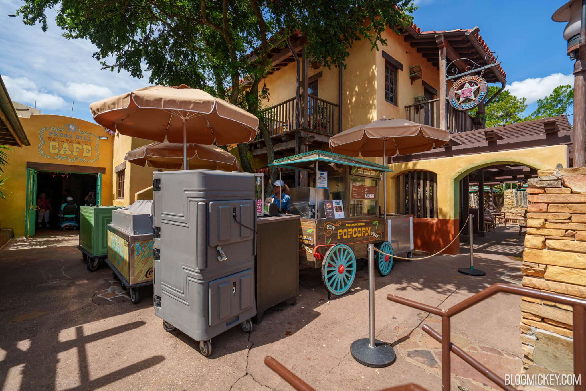 A wooden popcorn cart with turquoise spoked wheels and a painted popcorn sign, staffed by a cast member, positioned under tan umbrellas beside the yellow stucco walls and hanging sign of Pecos Bill Tall Tale Inn and Cafe.