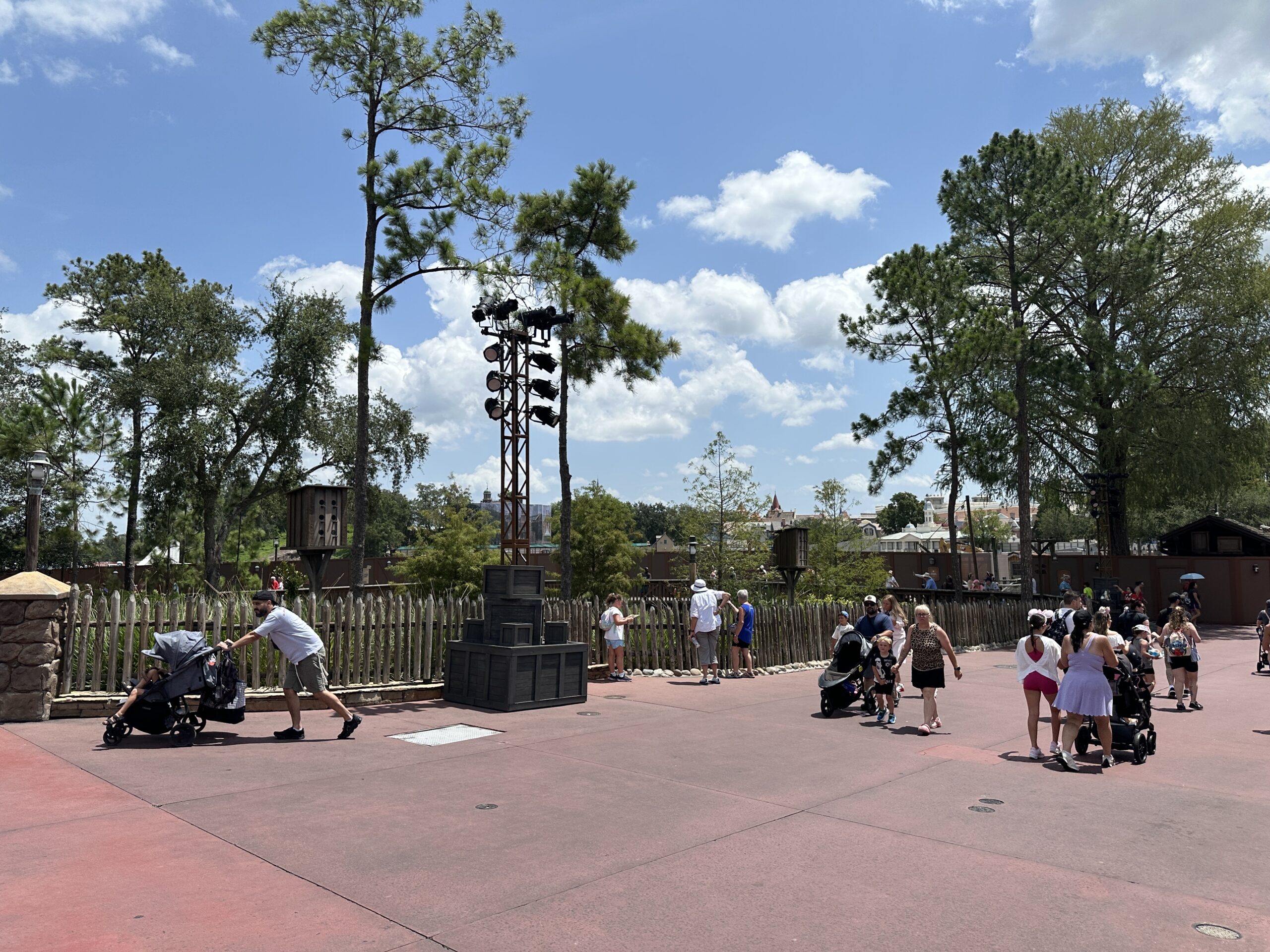 Guests walking along the wide reddish pavement of the Frontierland riverwalk area at Magic Kingdom on a sunny day, with a rustic wooden picket fence running along one side and brown construction walls visible beyond it, plus a tall lighting tower with black fixtures rising from a stack of grey wooden crates.