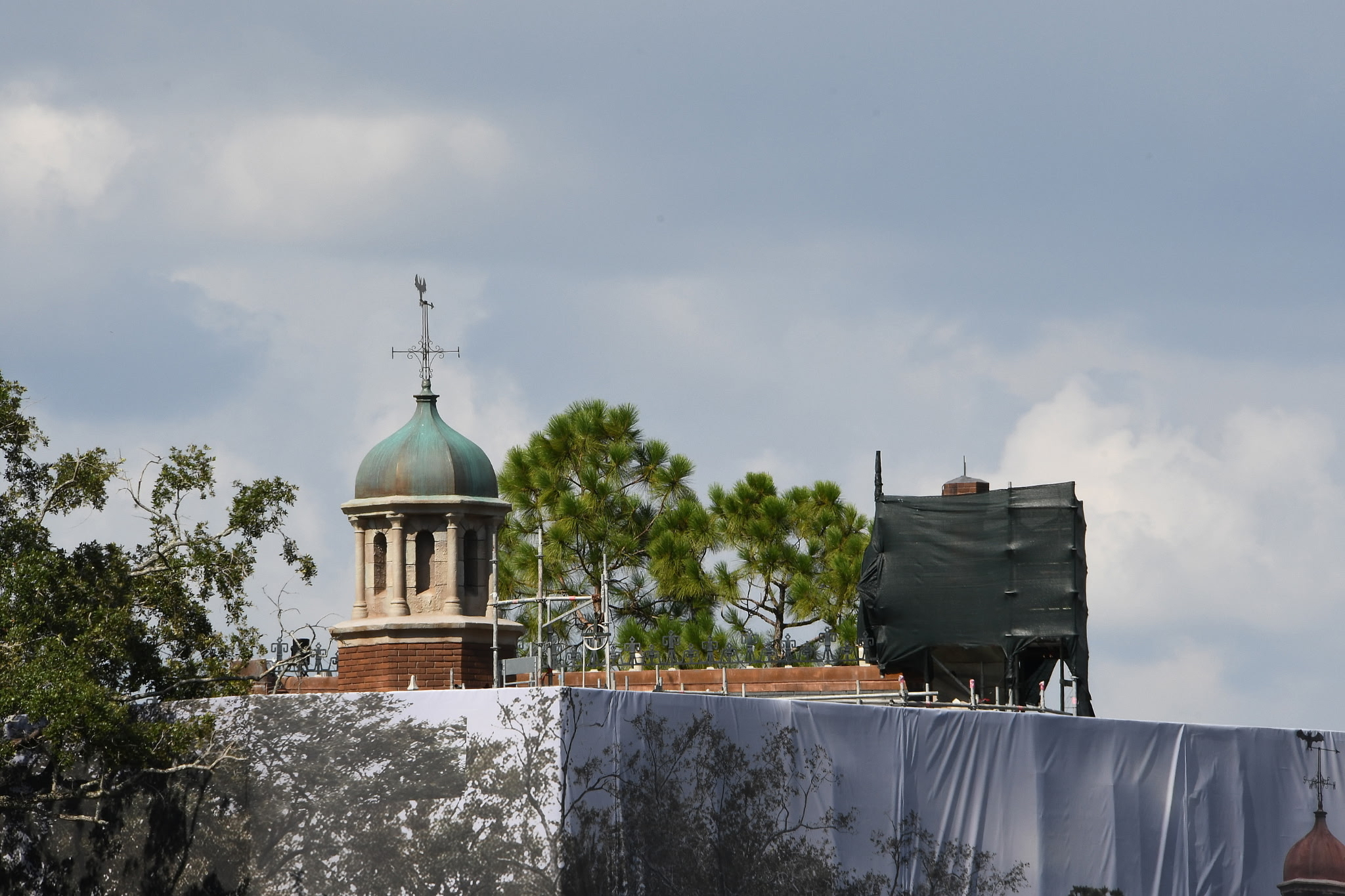 The Haunted Mansion's cupola with a weathervane, its dome a patina green above pale stone columns and a red brick base, with white construction scrim still covering the building below and dark green scrim over the chimneys at right