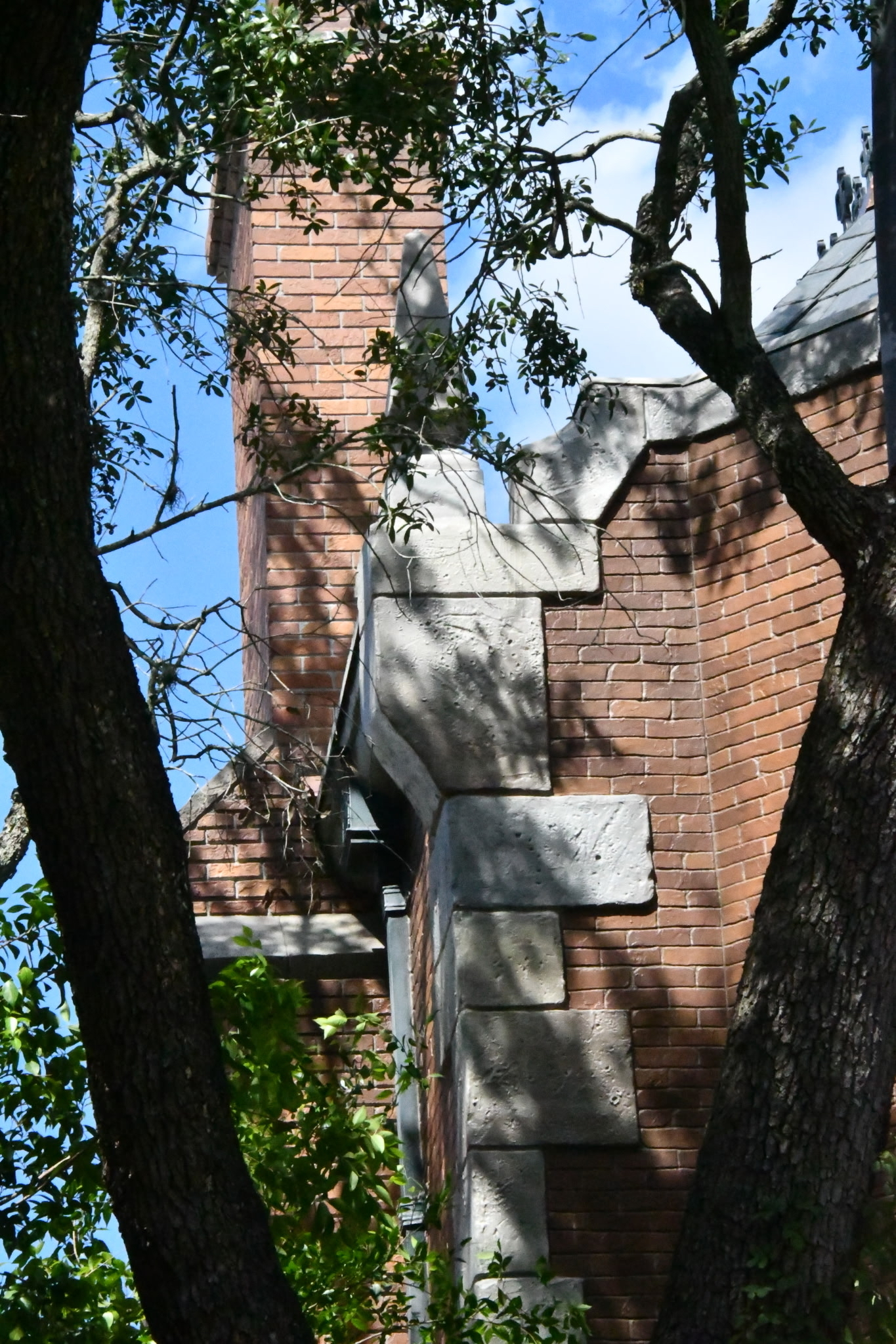 Light gray stone quoins stepping up the corner of the Haunted Mansion against darker red brick, with a brick chimney at left and gray roof shingles visible at upper right, framed by oak branches