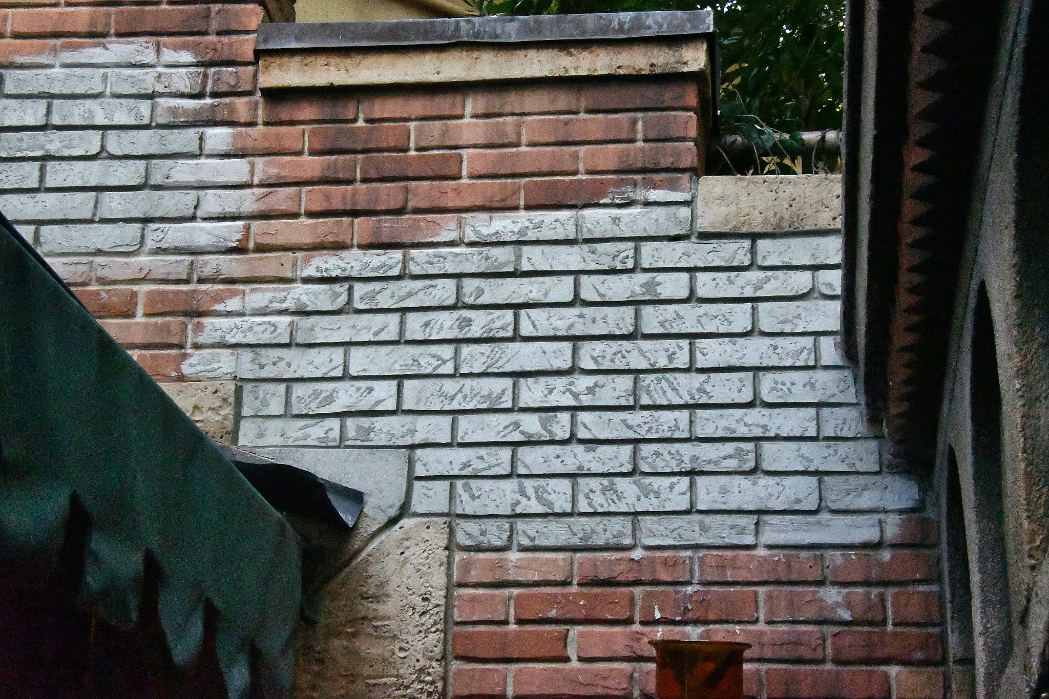 A brick wall where roughly half the bricks have been coated in patchy gray-white whitewash and the rest are still bare red brick, with a green tarp at the left edge
