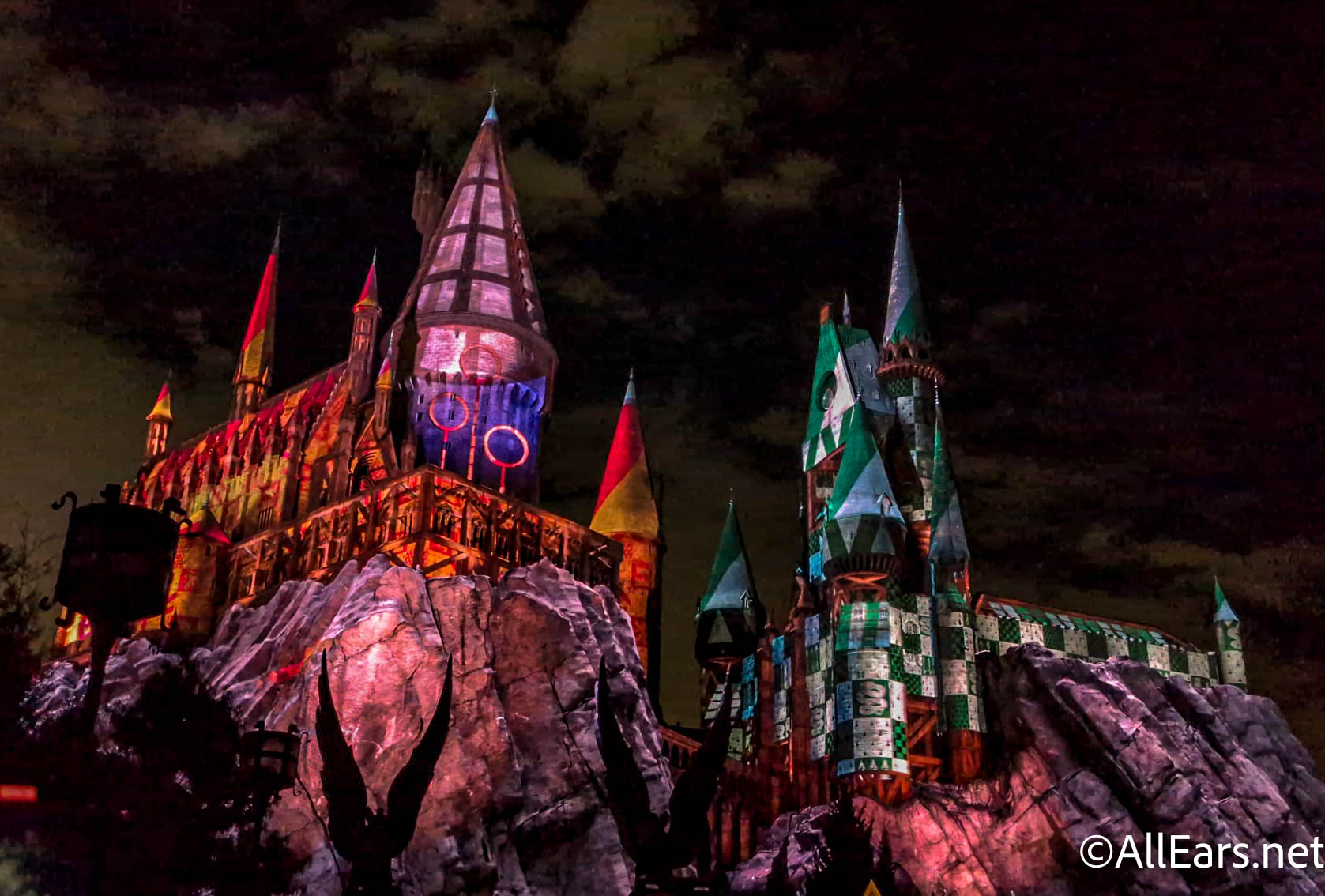 Hogwarts Castle at night washed in projection mapping, with the left towers lit in Gryffindor red and gold and the right towers in Slytherin green and silver