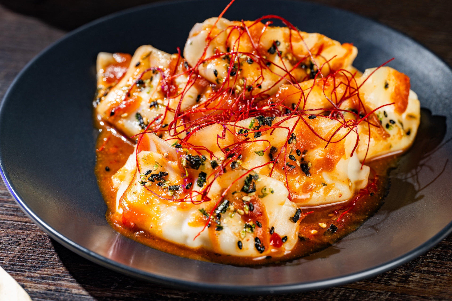 A black bowl of chicken dumplings coated in a glossy red miso gochujang sauce, topped with black and green furikake seasoning and a tangle of fine red chili threads, sitting on a dark wooden table.