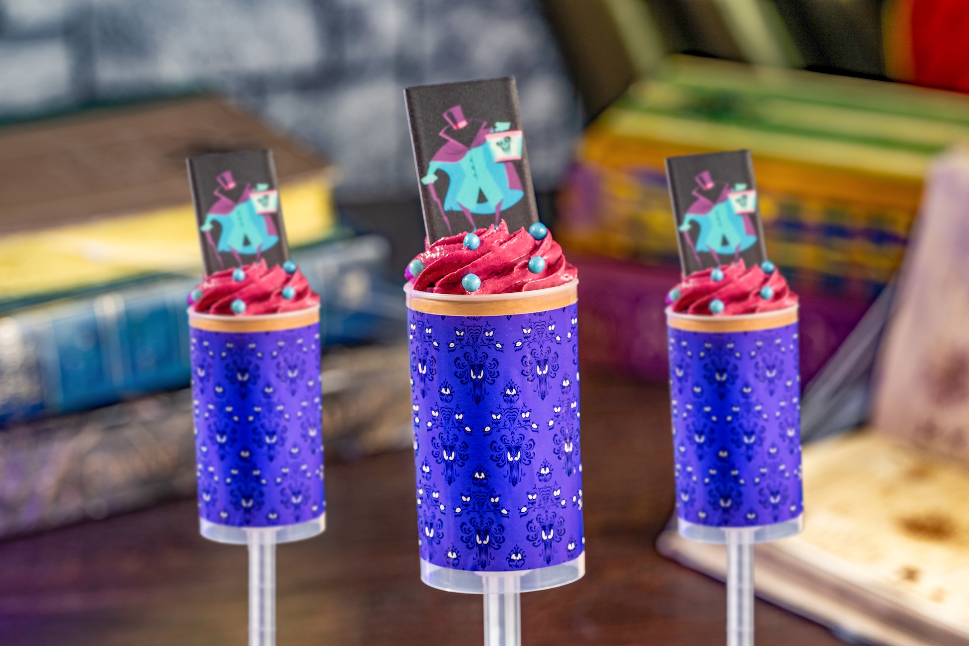 Three dessert push-pops in clear tubes wrapped in the purple Haunted Mansion wallpaper pattern, each topped with a swirl of magenta mousse, blue candy pearls and a flat chocolate piece printed with the Hatbox Ghost holding his hatbox.