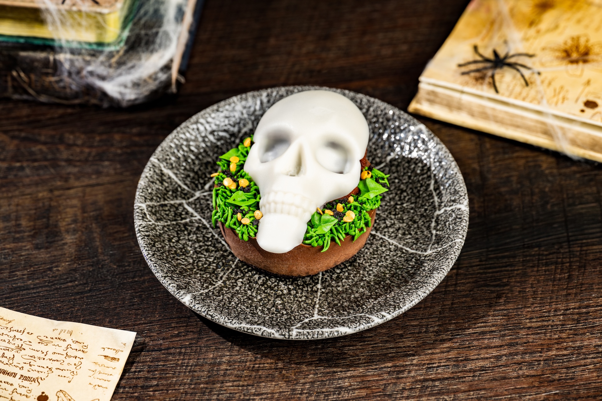 A round chocolate dessert topped with a glossy white skull resting on green piped buttercream and orange candy pieces, plated on a speckled grey dish beside cobweb-draped books and an aged parchment scroll.