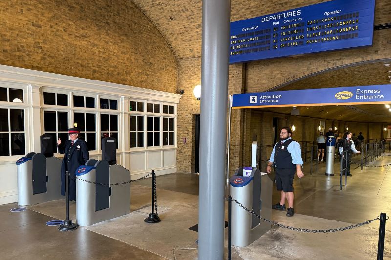 Team members standing beside stainless steel ticket validation posts at the Hogwarts Express entrance inside King's Cross Station at Universal Studios Florida, under a brick vaulted ceiling with a blue departures board and blue Attraction Entrance and Express Entrance signs.