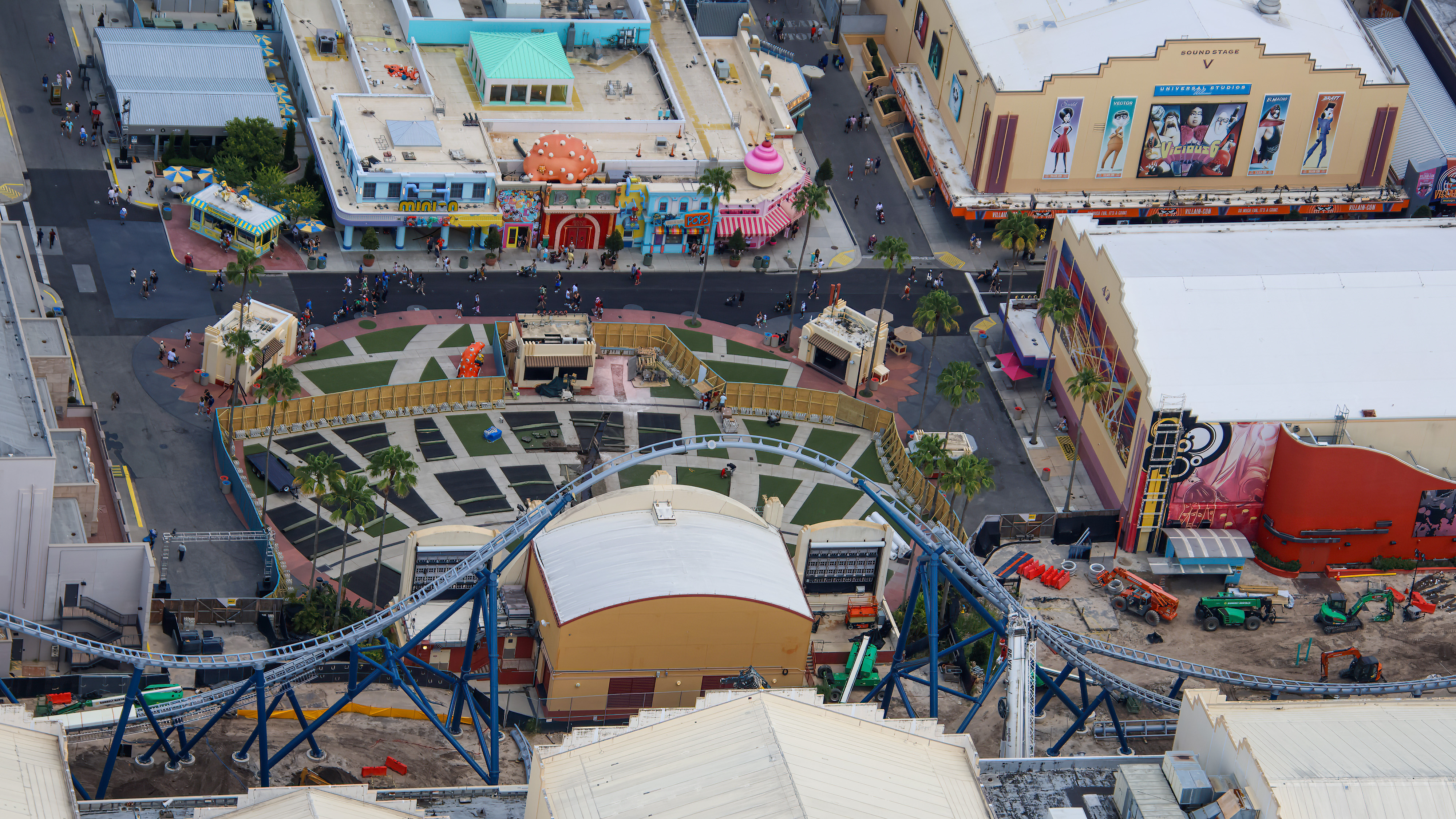 Aerial view looking down on the Music Plaza lawn at Universal Studios Florida, showing the checkerboard of green turf squares with roughly half stripped to dark base panels, construction fencing snaking through the plaza, palm trees, and the colorful facades of Minion Land across the street.