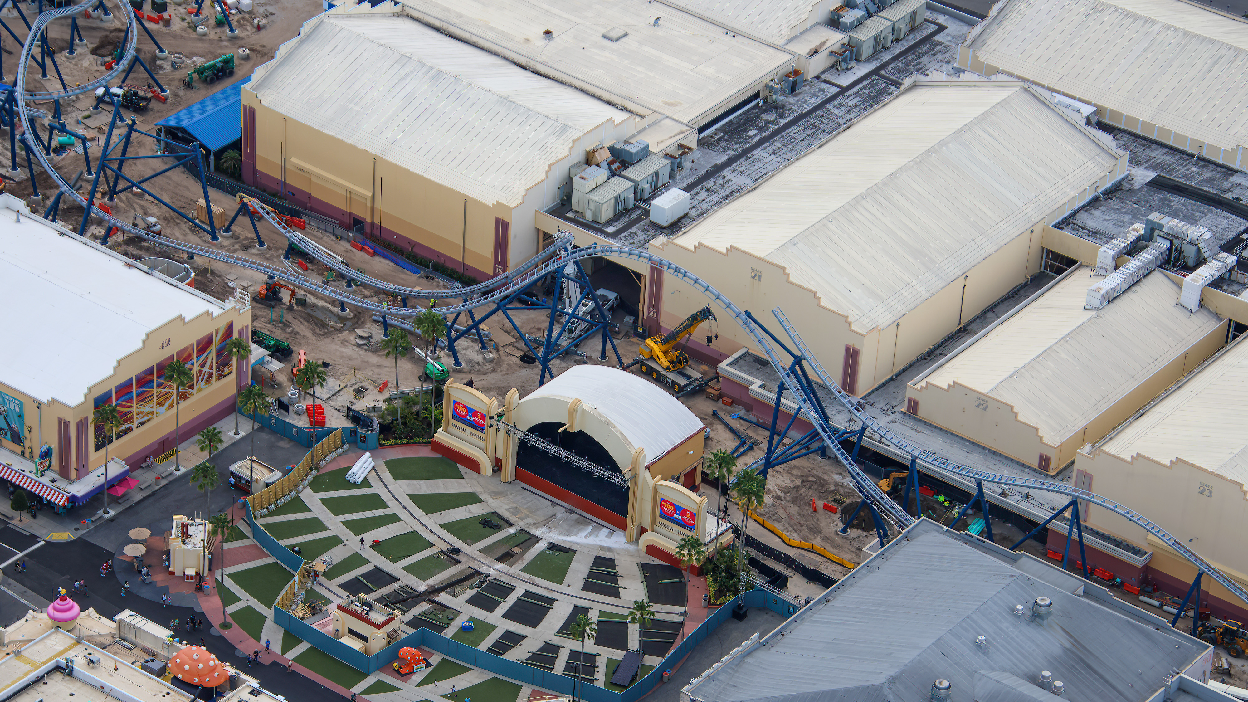 Aerial view of the area behind Music Plaza Stage at Universal Studios Florida, showing Hollywood Drift coaster track dipping directly behind the stage structure, a yellow mobile crane at work, blue support columns, and the partially stripped concert lawn in the foreground.