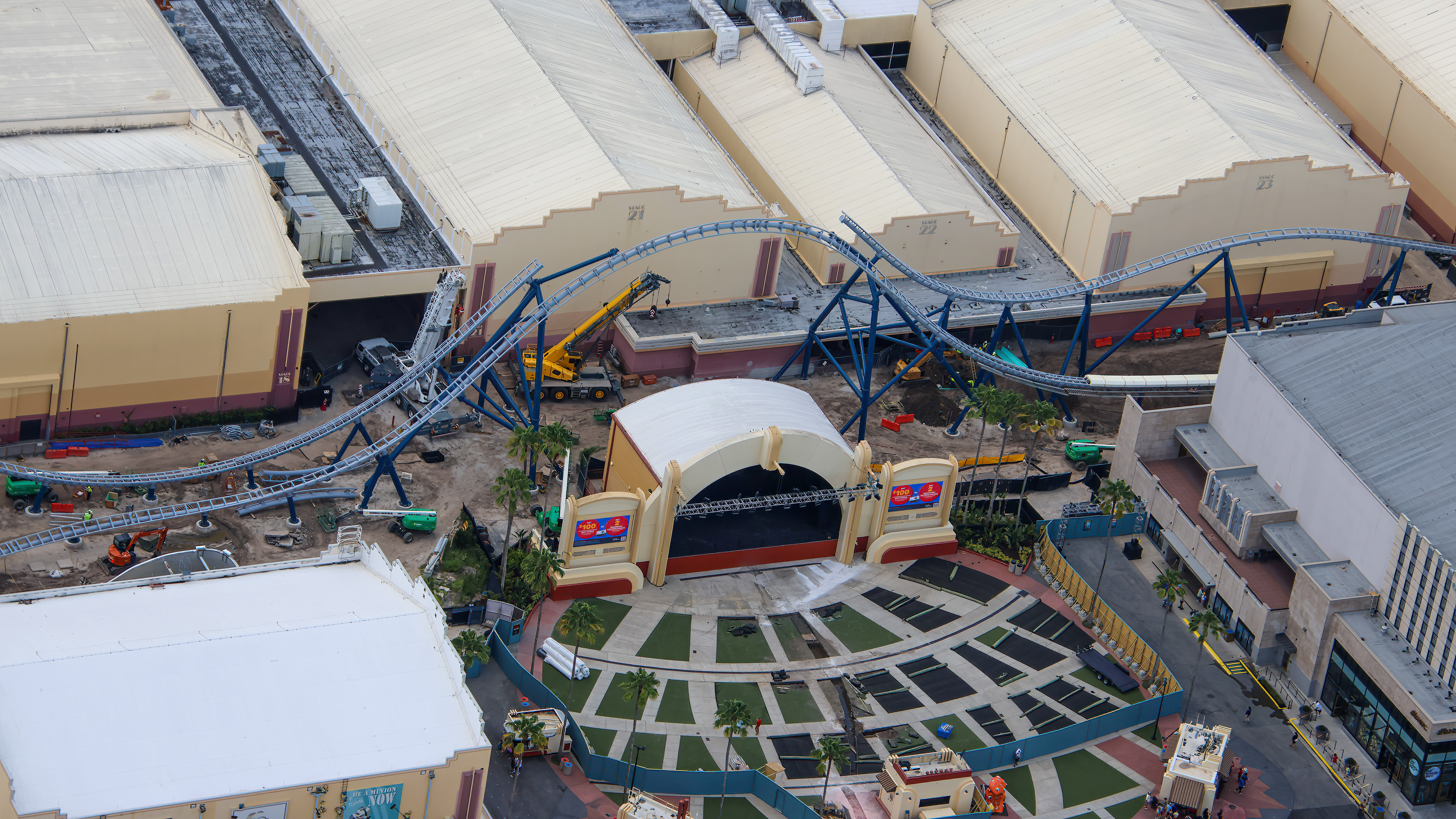Aerial view of Fast & Furious: Hollywood Drift construction at Universal Studios Florida, with gray coaster track arcing over the corridor beside Soundstages 21 and 22, dipping low behind the arched roof of Music Plaza Stage, and two mobile cranes working the section.