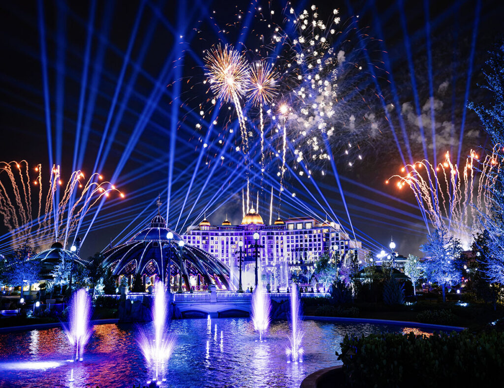 Fireworks and blue searchlight beams above illuminated fountains in Celestial Park, with the domed Constellation Carousel at left and the Universal Helios Grand Hotel lit behind