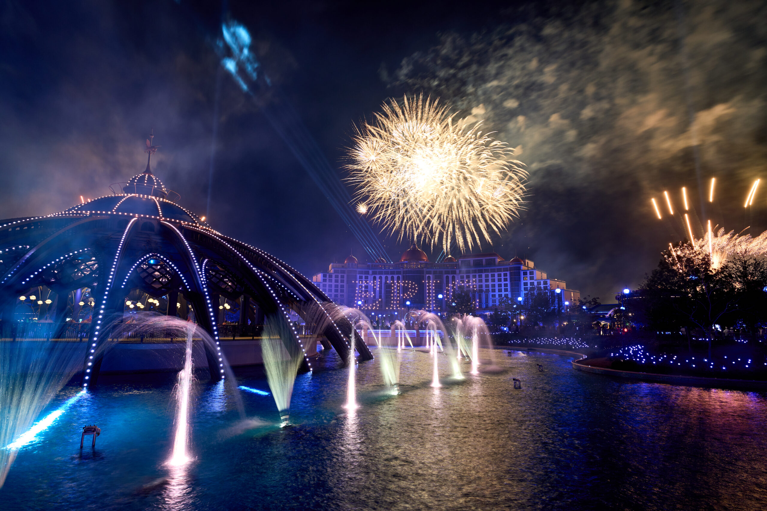 A wide night view of the Celestial Park lagoon with lit fountains, the domed Constellation Carousel at left, a golden firework burst overhead and the Universal Helios Grand Hotel lit behind