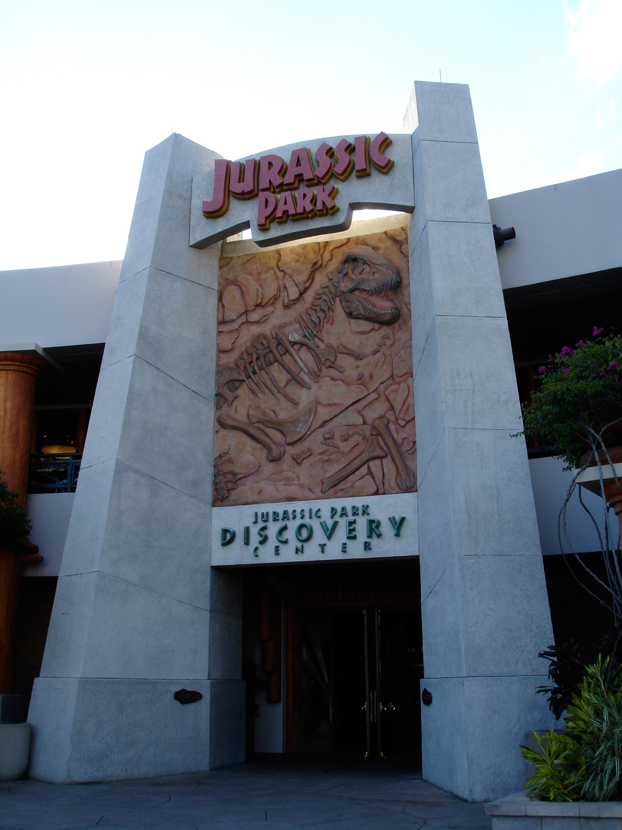 The Jurassic Park Discovery Center entrance, a tall grey stone facade with a Jurassic Park sign above a sculpted Tyrannosaurus fossil relief