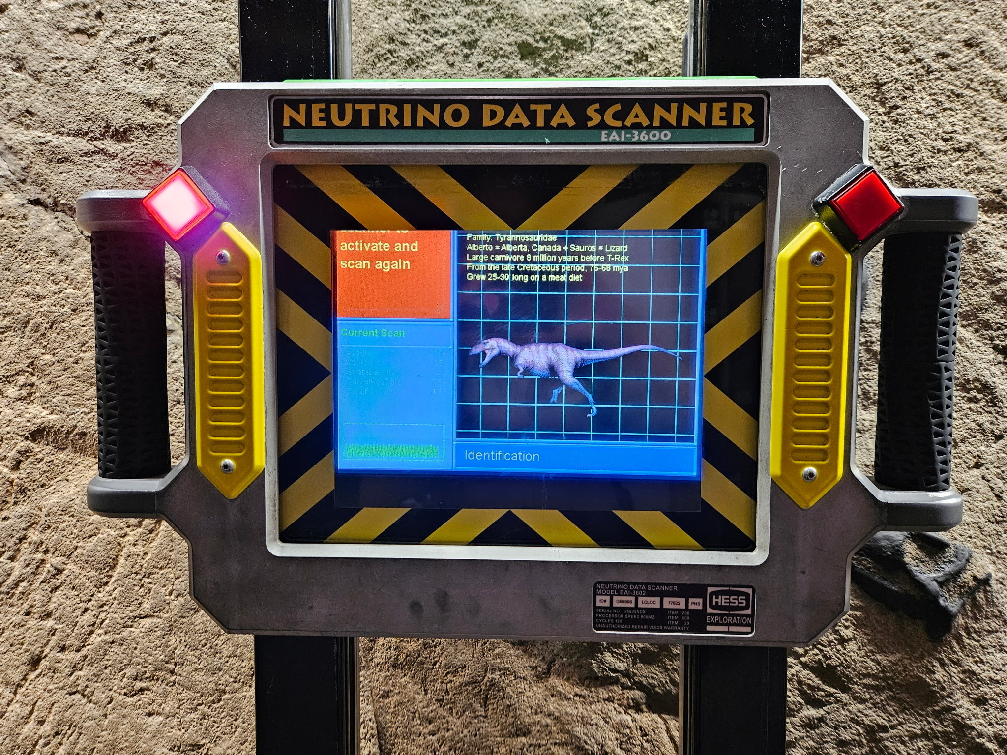 A Neutrino Data Scanner prop mounted on black vertical poles against a rock wall, its screen showing a blue wireframe scan of a dinosaur alongside identification text