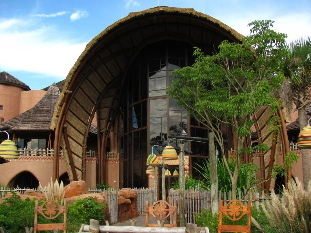 A tall thatched arch over a glass window wall at Kidani Village, with striped gourd lanterns hanging nearby and a rustic wooden fence along a garden walkway in the foreground