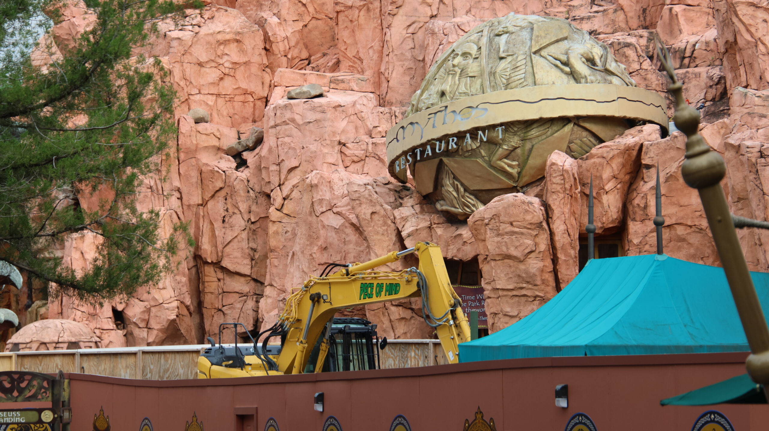 A yellow excavator behind a wooden construction wall just below the golden globe sign of Mythos Restaurant at Islands of Adventure.