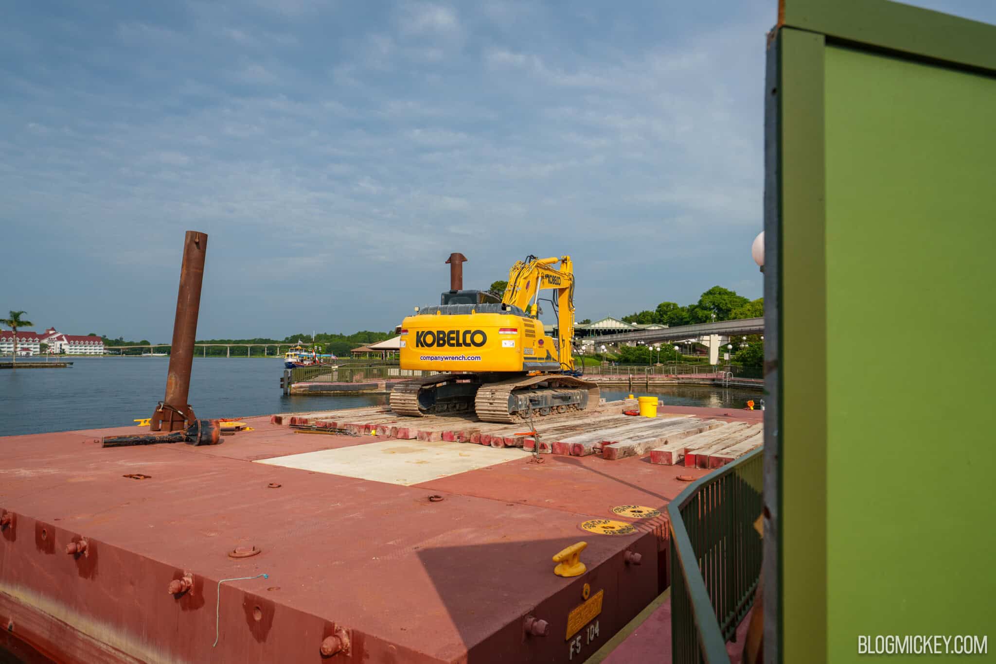A close view of the barge deck showing a yellow Kobelco excavator, stacked wooden timbers and an upright steel pile.