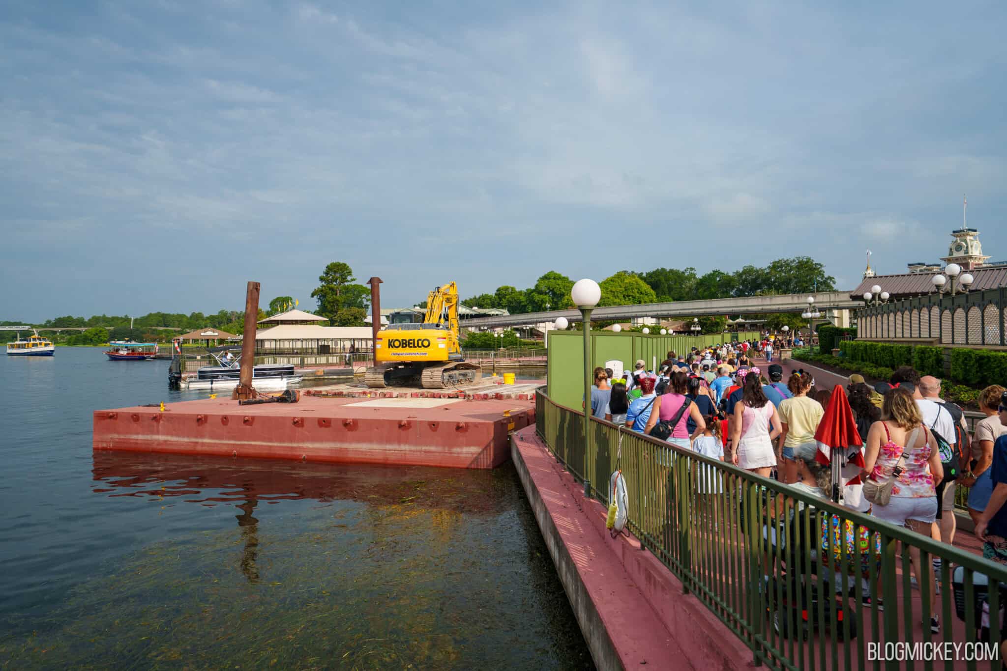 A red construction barge holding a yellow excavator floats in the Seven Seas Lagoon beside the guest walkway to Magic Kingdom.
