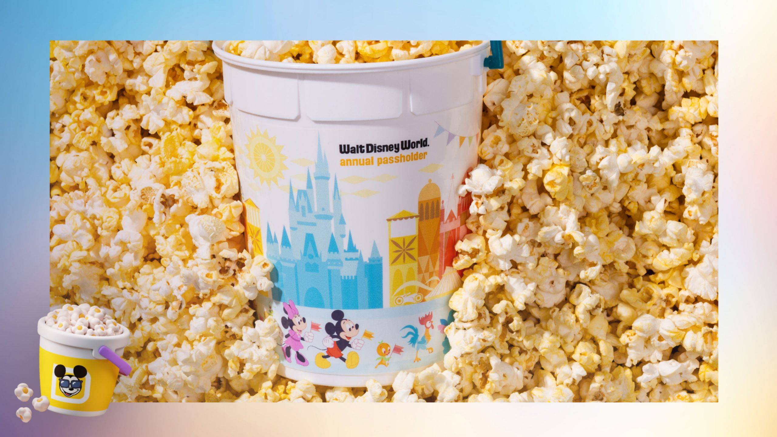 A white Walt Disney World annual passholder popcorn bucket sitting in a bed of popcorn, printed with Cinderella Castle, stylized icons from the four theme parks and a parade of characters including Minnie, Mickey and the Orange Bird