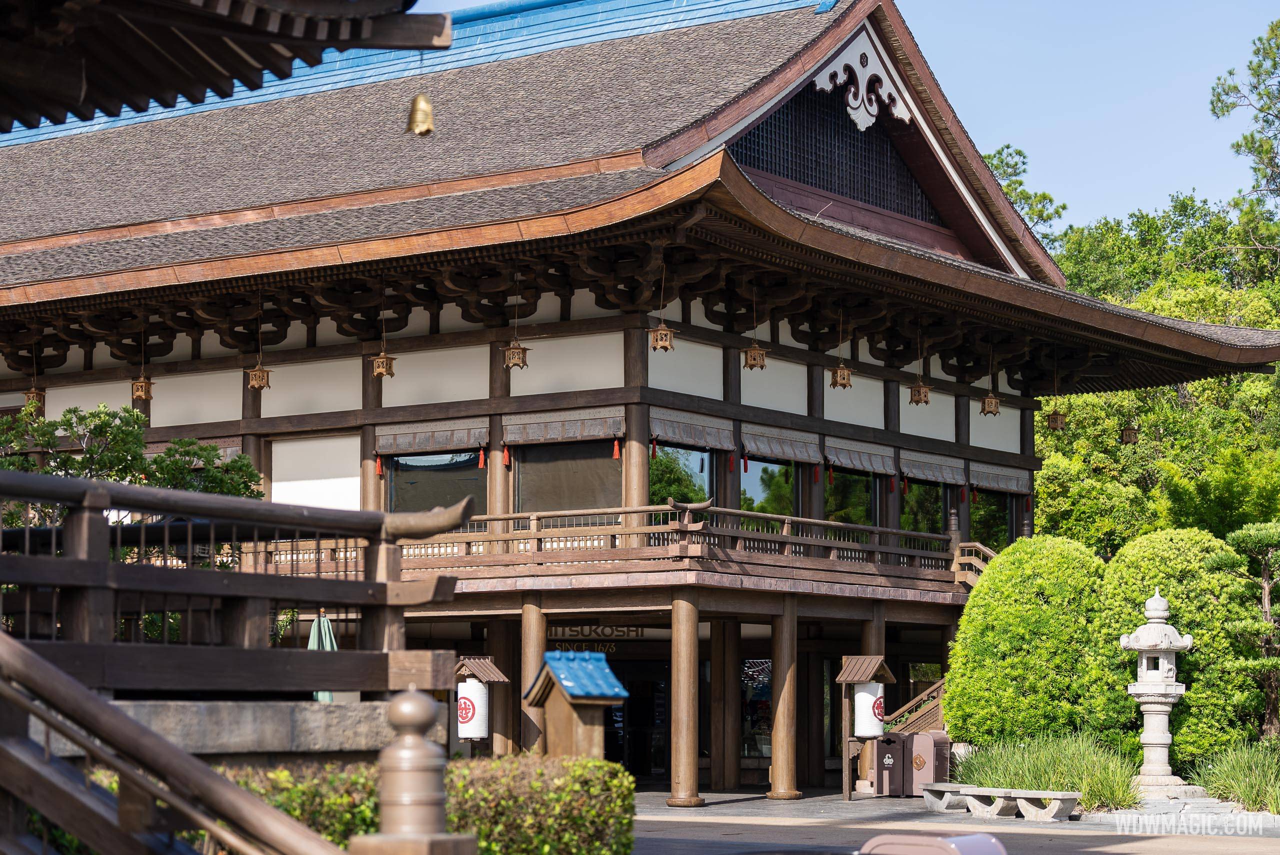Daytime exterior of the large wooden Mitsukoshi building in EPCOT Japan Pavilion, with hanging lanterns under deep eaves, a stone lantern at right and blue sheeting covering part of the upper roof