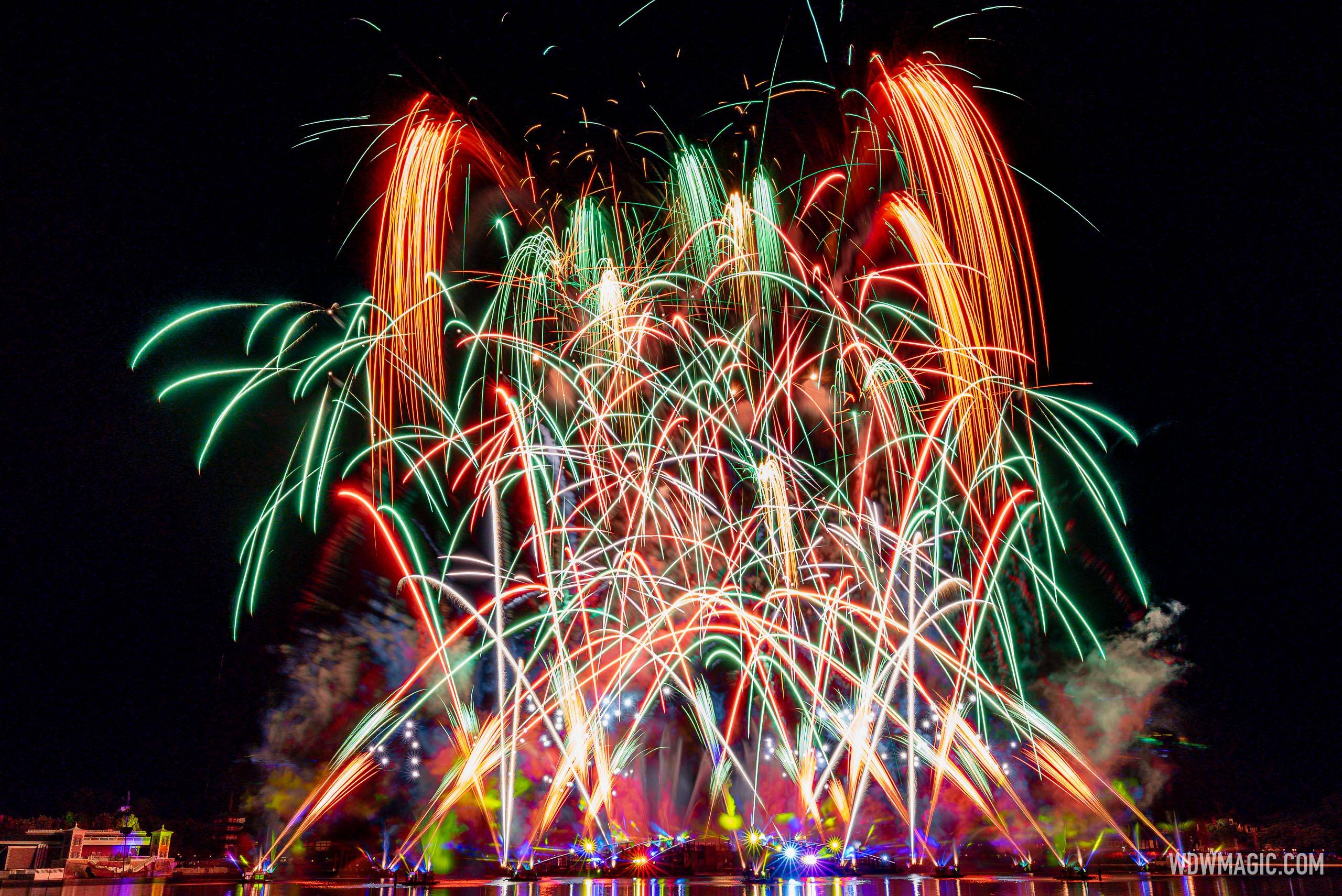 Red, green and gold fireworks bursting in a wide fan over World Showcase Lagoon at EPCOT, with lit barges reflected in the water below