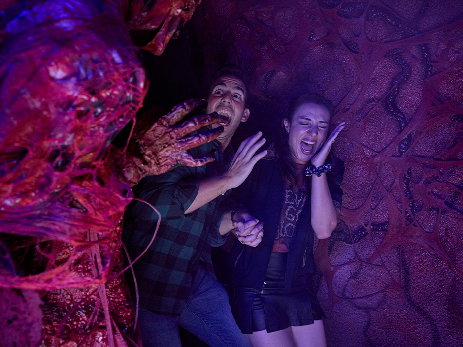 Two guests holding hands and screaming inside a dark red and purple corridor with fleshy organic walls, as a clawed hand reaches toward them from the left