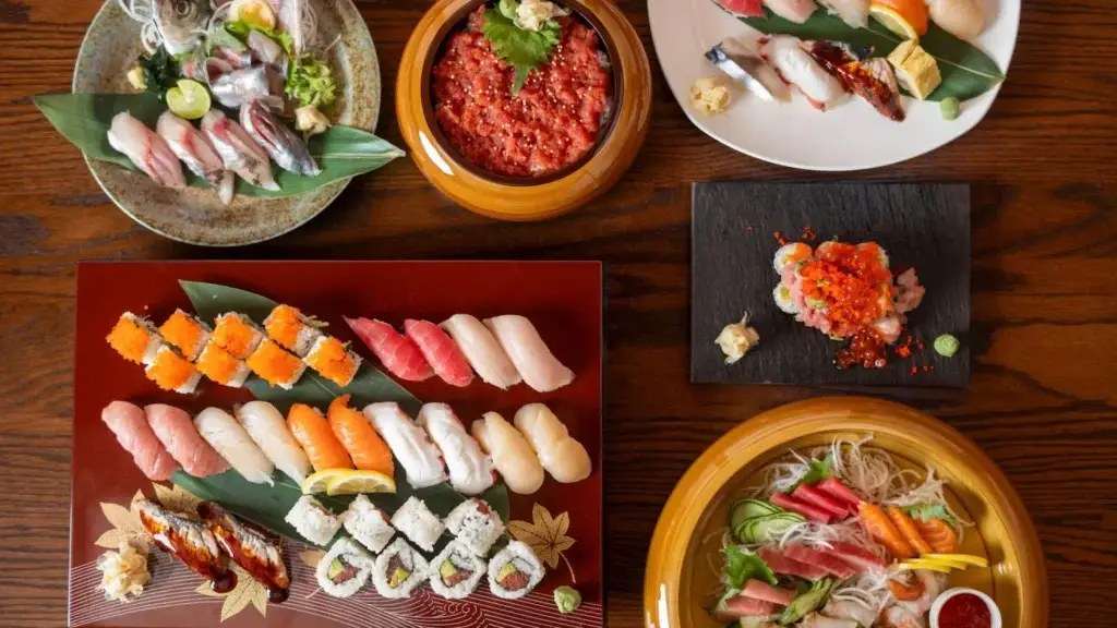 Overhead view of sashimi, nigiri, sushi rolls, a chopped tuna bowl topped with roe and a noodle and sashimi bowl arranged on a wood table