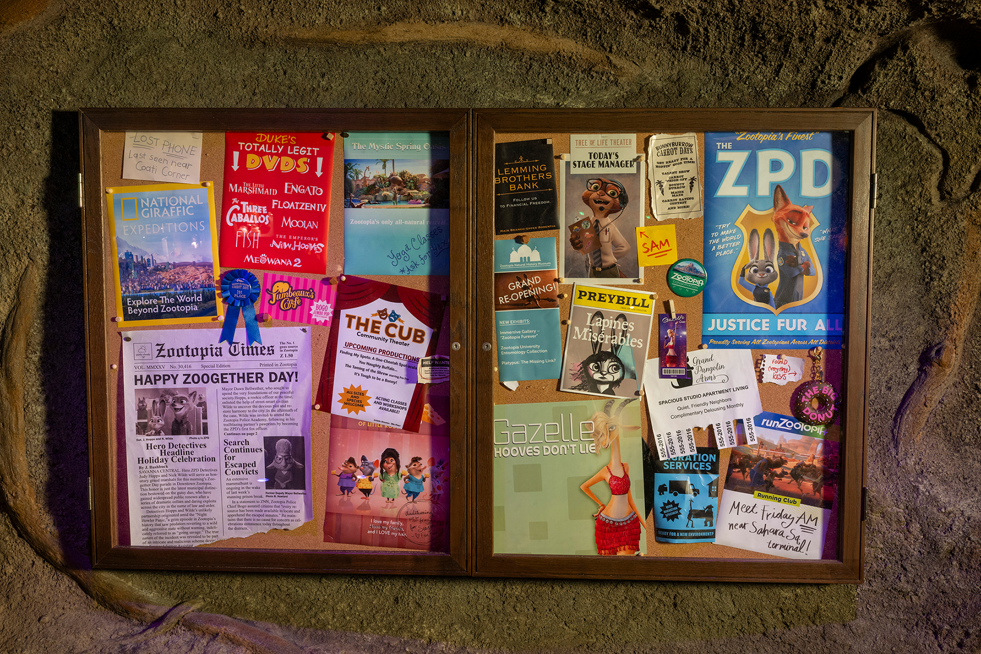 A wooden bulletin board case set into a rock wall, filled with Zootopia in-universe posters, flyers and a Zootopia Times newspaper with the headline Happy Zoogether Day