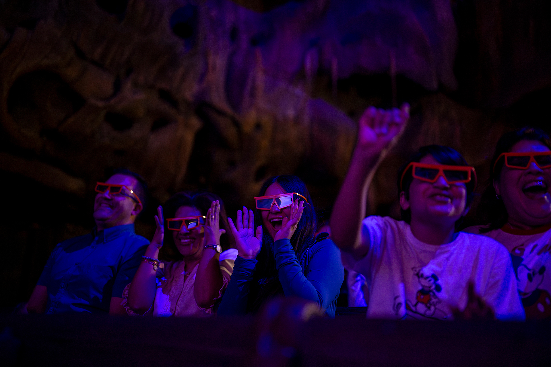 An audience in the darkened Tree of Life Theater wearing red-framed Carrot Vision glasses, laughing and reaching upward under blue and purple lighting