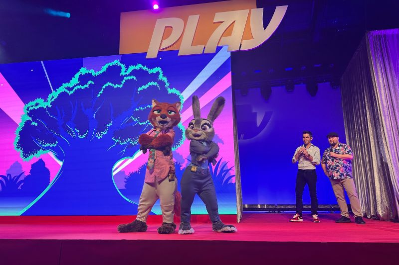 Costumed Nick Wilde and Judy Hopps characters standing on a stage in front of a bright blue and pink stylized tree graphic, with two presenters to the right