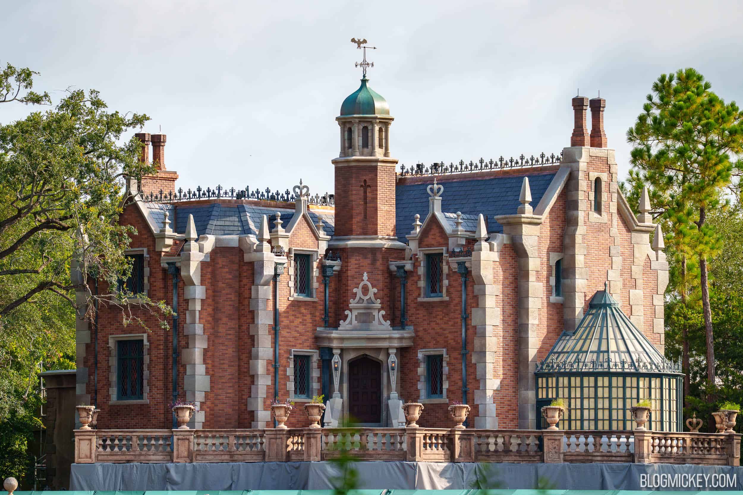 The Haunted Mansion facade at Magic Kingdom fully revealed, showing dark reddish brick, gray stone trim, a dome finial with a green patina, a dark roof and the glass greenhouse to the right