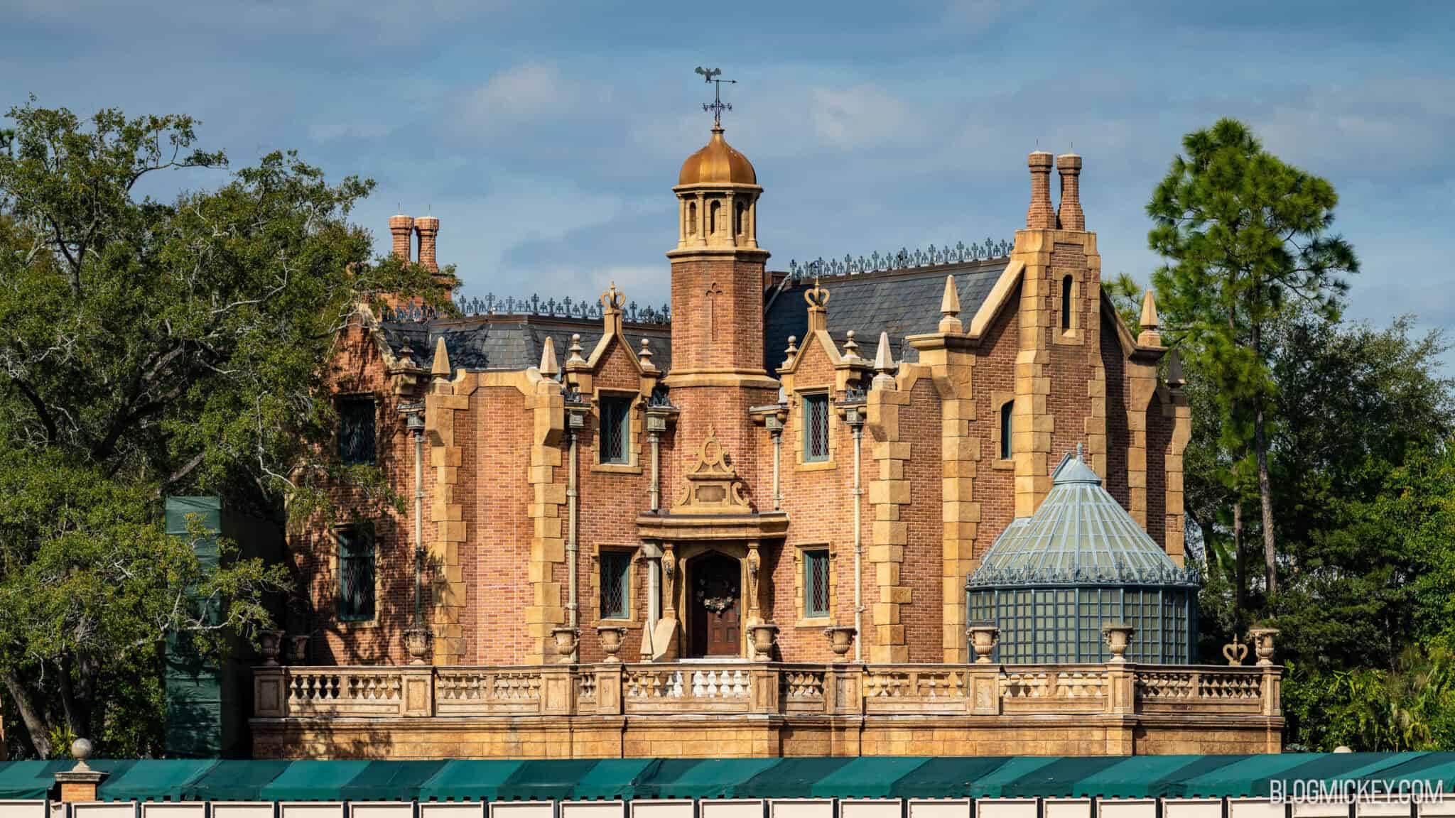 The Haunted Mansion in January 2026 before the repaint, with a gold dome finial above the entrance tower, and brick and stone in similar tan tones, behind a green construction wall