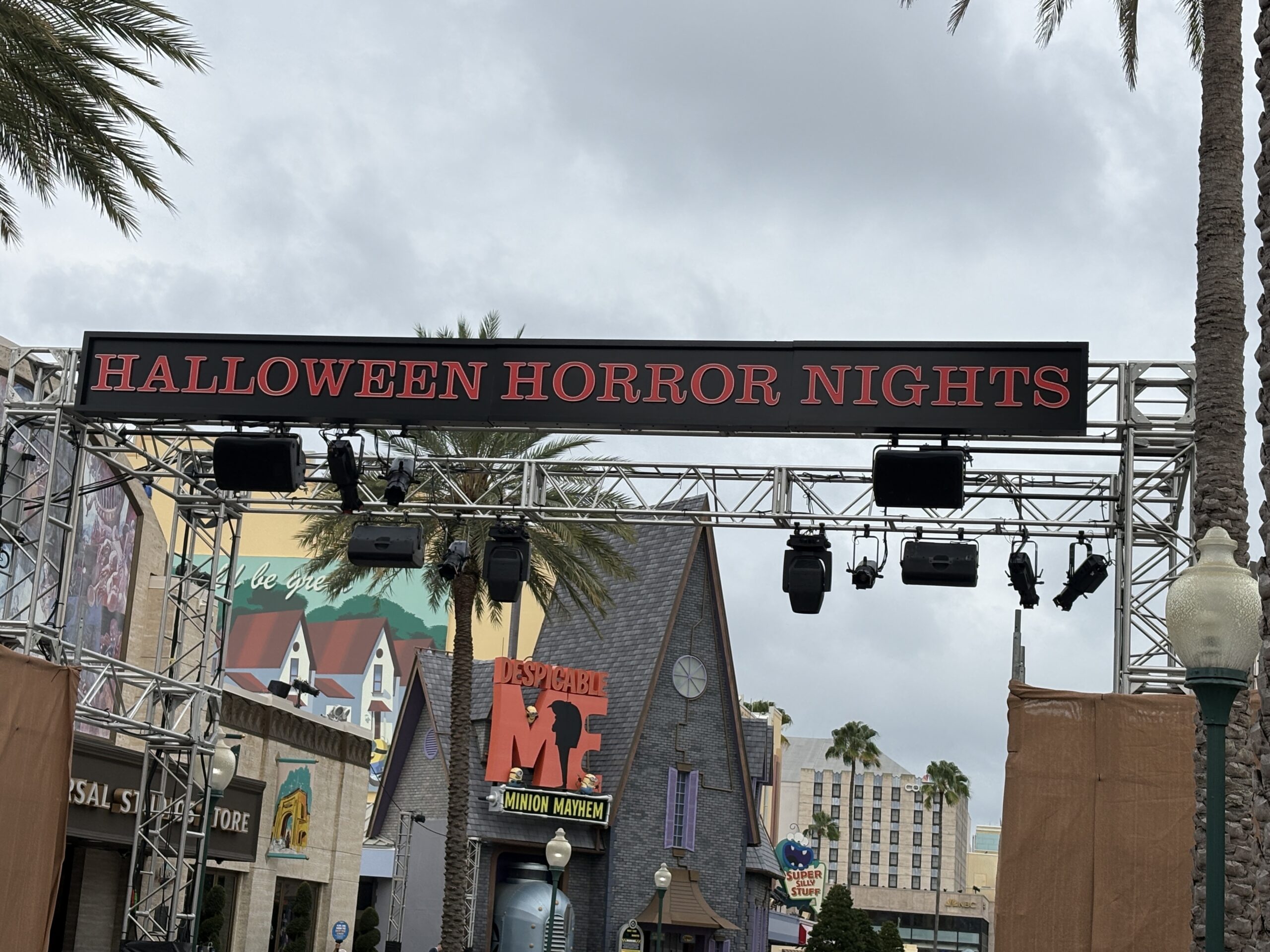 A long black sign reading HALLOWEEN HORROR NIGHTS in red serif letters, mounted on a metal truss with stage lights and speakers hanging below it, with the Despicable Me Minion Mayhem facade and palm trees behind