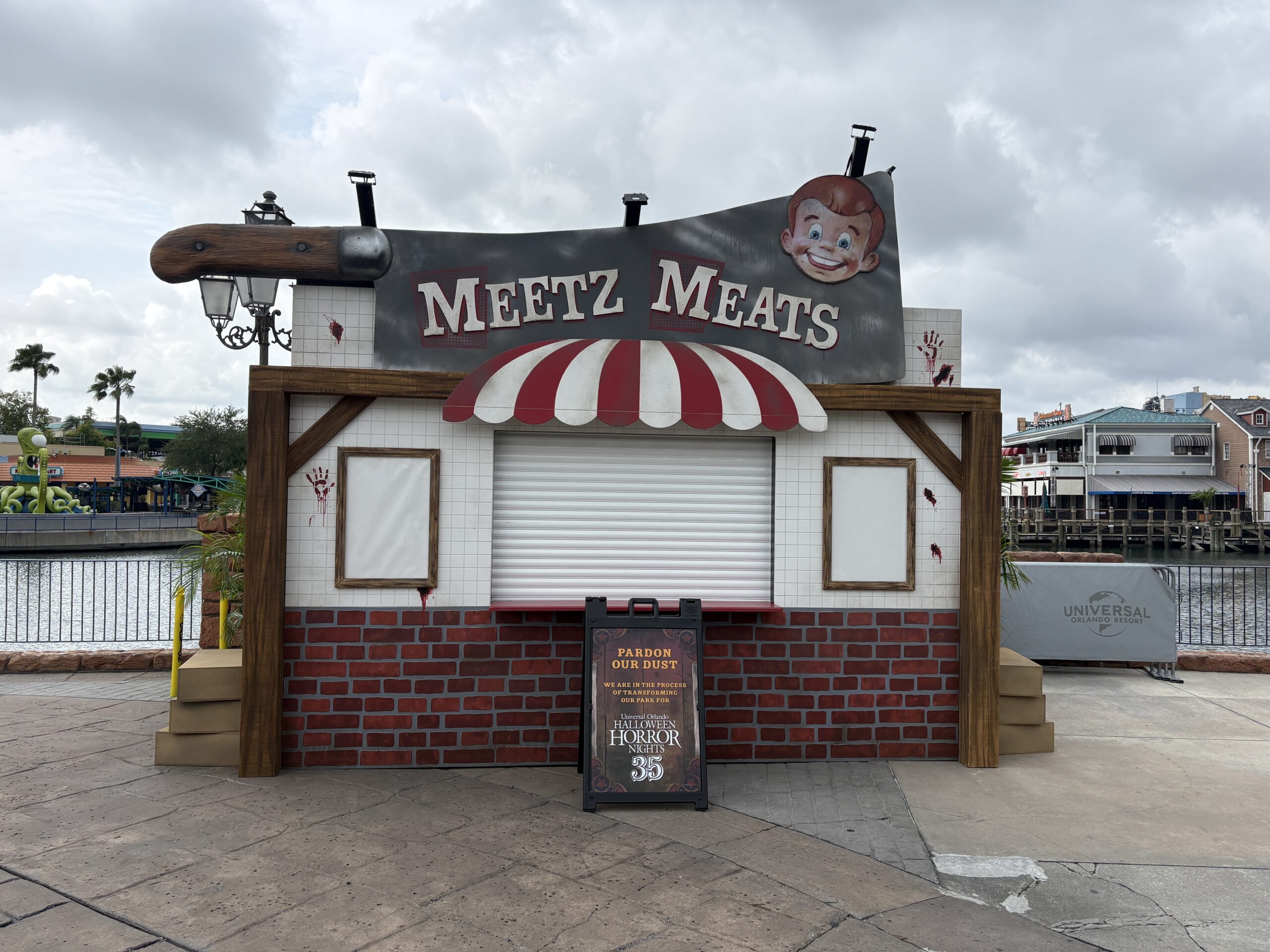 A food booth shaped like a butcher shop with a giant meat cleaver forming its sign reading MEETZ MEATS, a grinning cartoon mascot face on the blade, red and white striped awning, white tile, red brick and bloody handprints