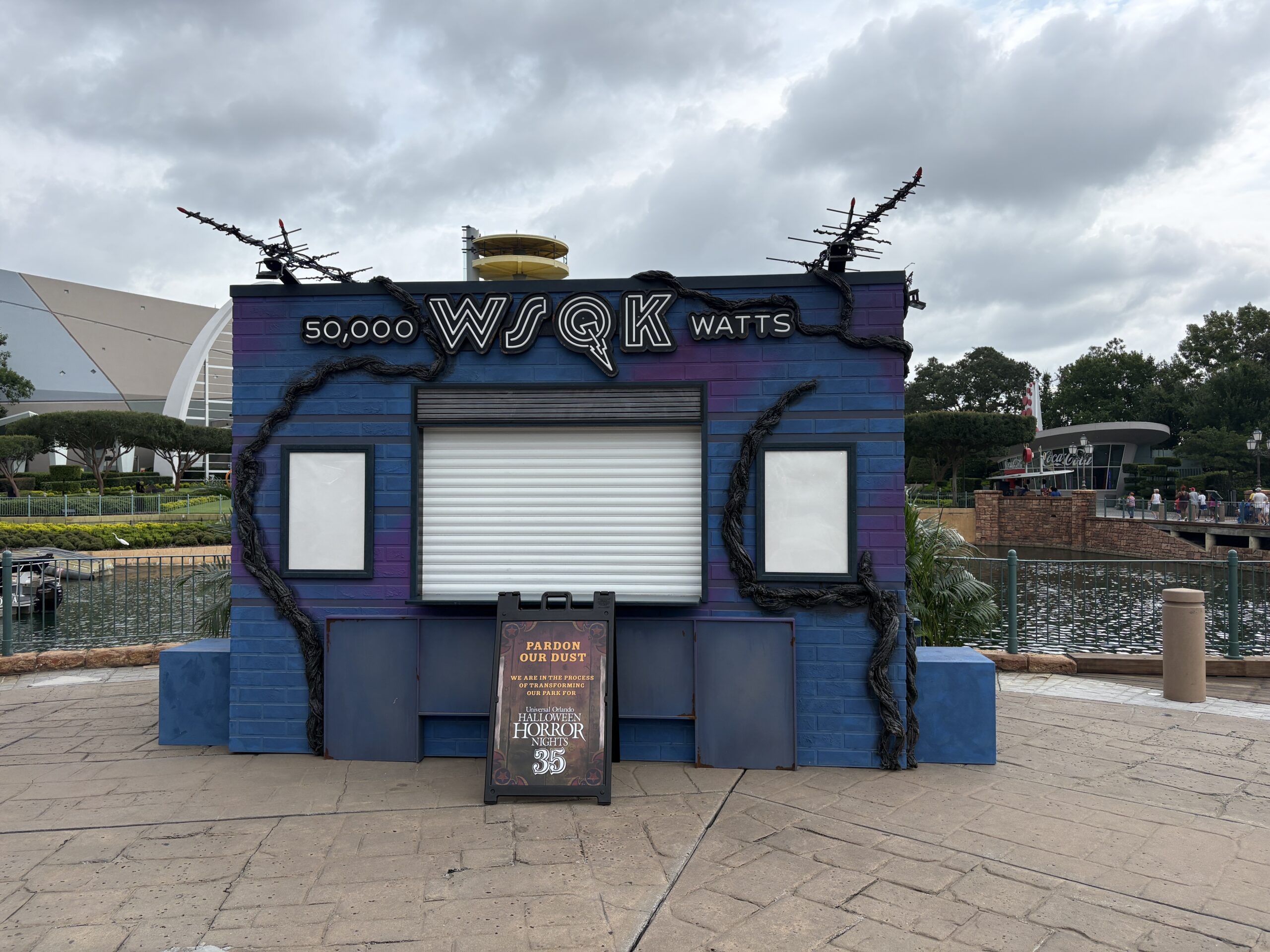 A blue and purple food booth built to look like a brick radio station building, with a sign reading 50,000 WSQK WATTS, antenna arrays on the roof, black vines across the walls, and a closed roll-up shutter