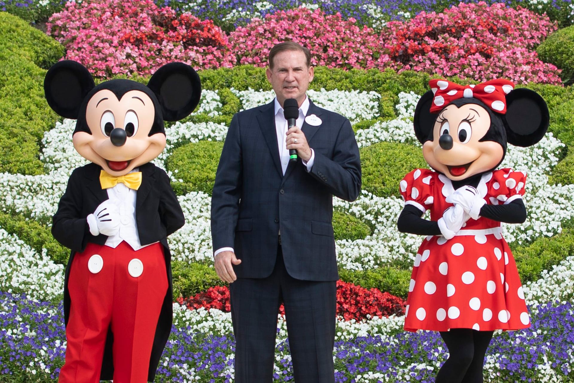 A man in a dark suit holding a microphone stands between Mickey Mouse and Minnie Mouse in front of a large flower bed