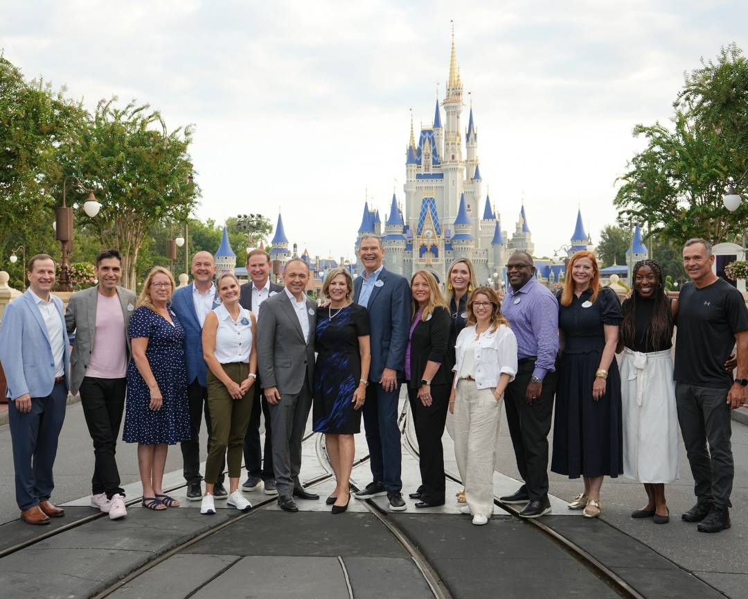 About sixteen people in a mix of business and casual attire stand together on the Main Street trolley tracks with Cinderella Castle behind them