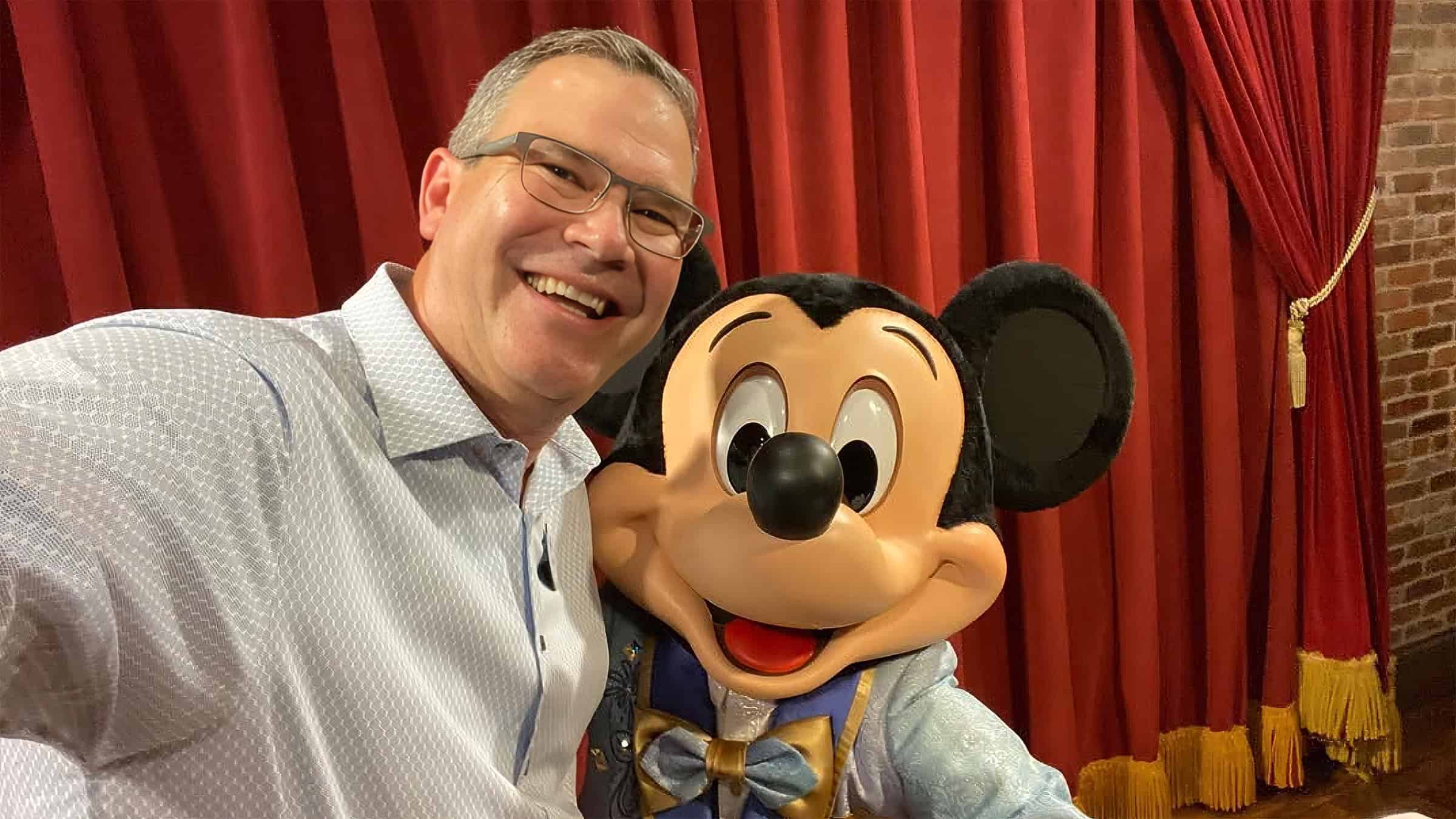 Jeff Vahle, in a light blue patterned shirt and wire rim glasses, smiles cheek to cheek with Mickey Mouse in a blue and gold costume in front of a red stage curtain